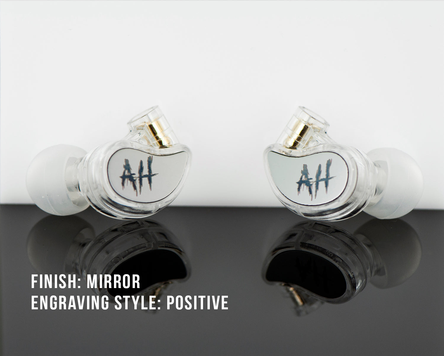 Two transparent heart-shaped earphones with the initials "ah" engraved in blue on a white background, reflecting on a shiny black surface. text: "finish: mirror engraving style: positive.