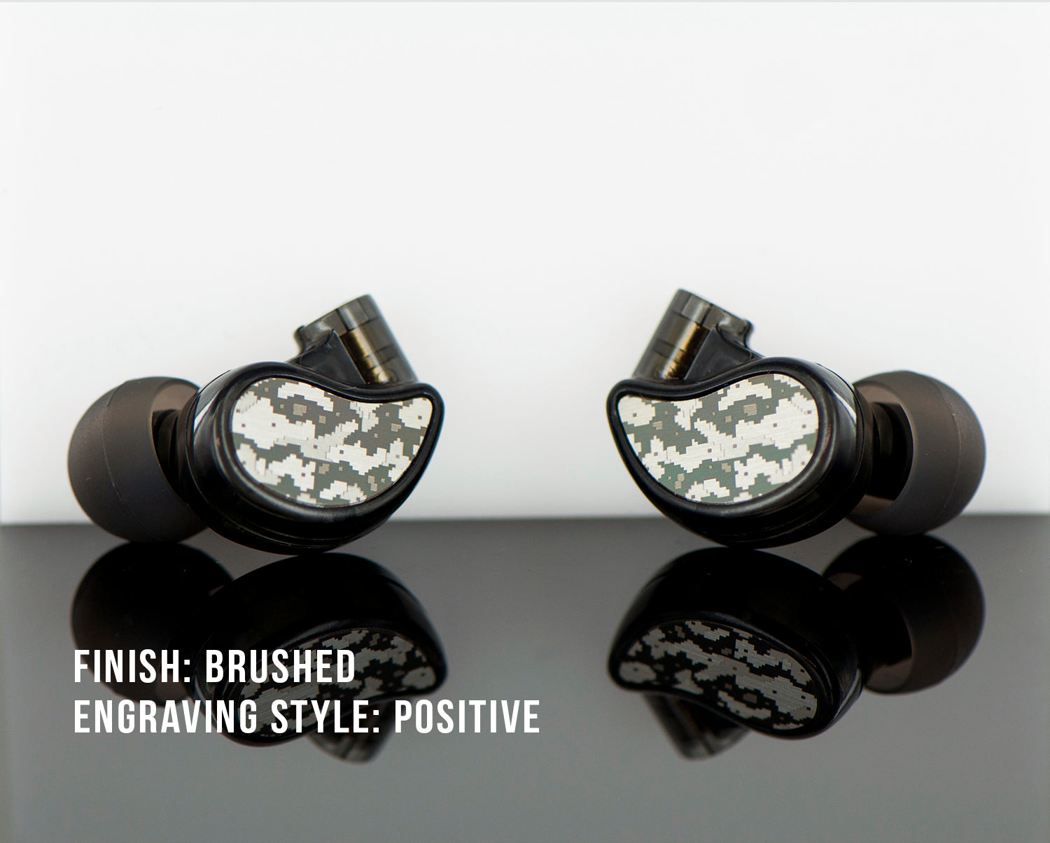 Two camouflage-patterned in-ear monitors displayed on a glossy surface with text describing them as having a brushed finish and positive engraving style.