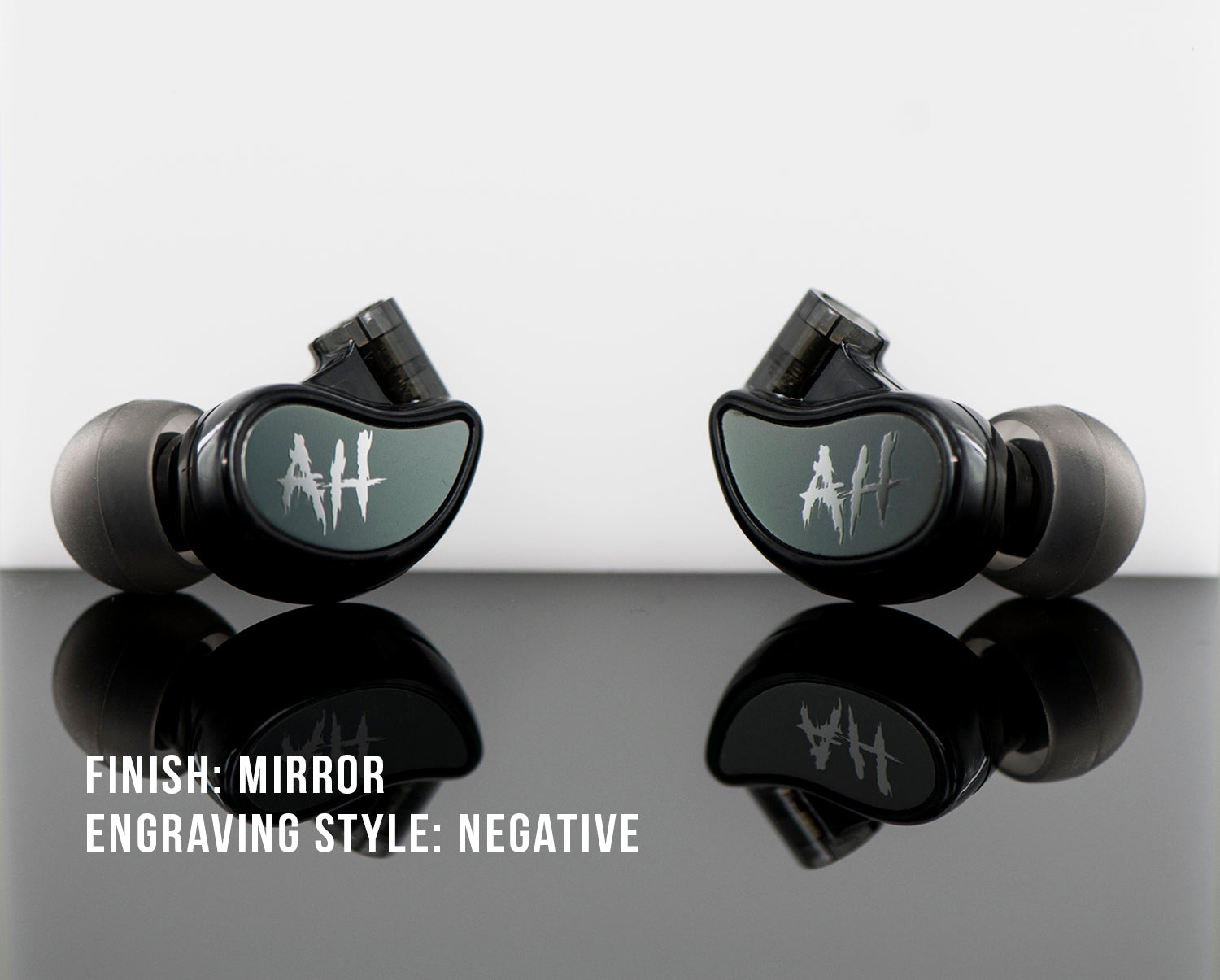 Two black in-ear headphones with a mirrored finish and a negative-style heart rate engraving, displayed on a reflective surface with the text "finish: mirror engraving style: negative.