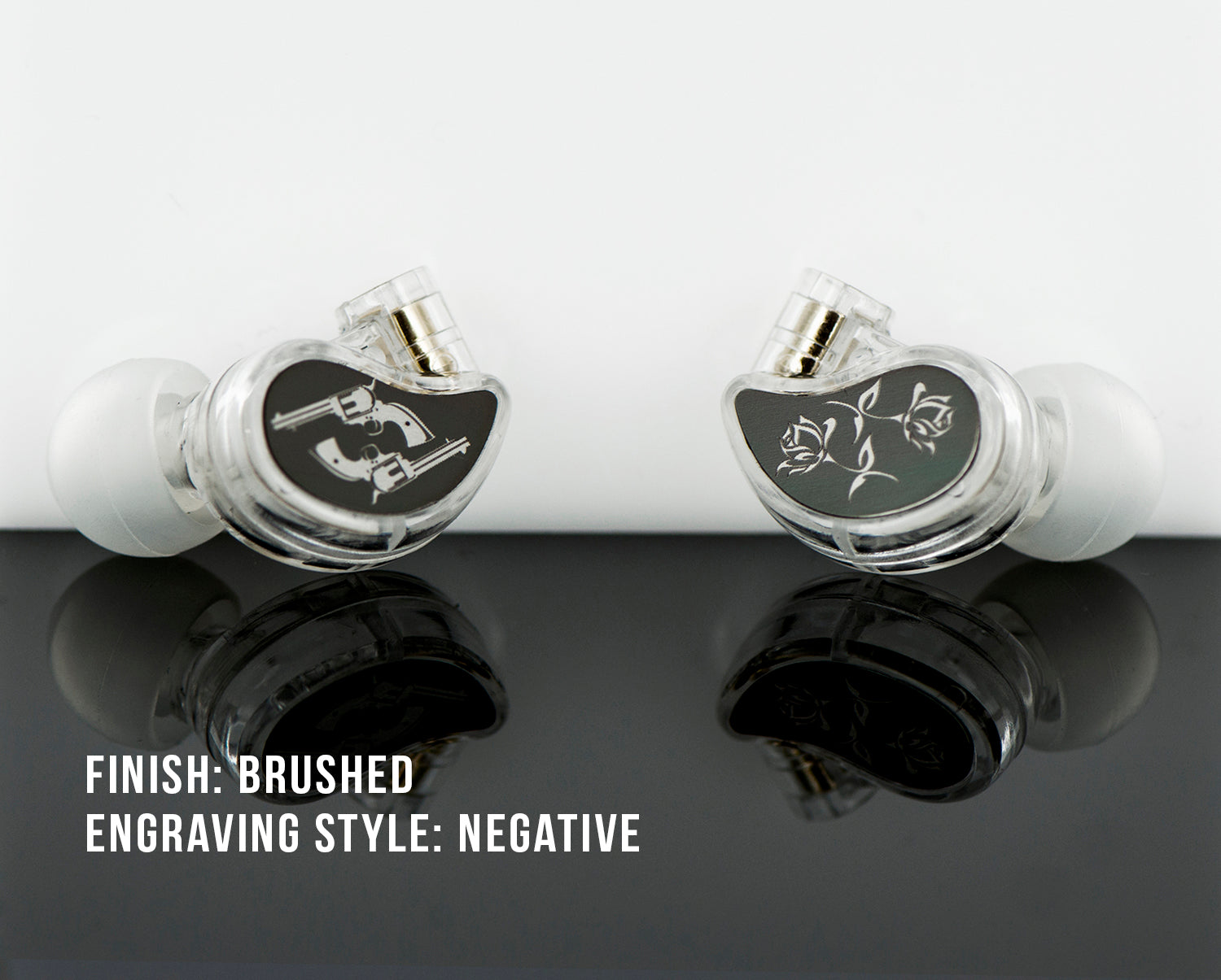 Two custom-engraved, clear in-ear monitors with brushed finish, displaying musical note and rose designs, reflected on a glossy black surface. text indicates "finish: brushed, engraving style: negative.