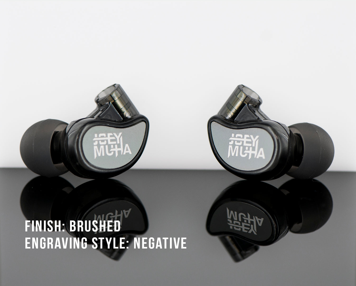 Two black in-ear monitors with "jh audio" branding, presented on a reflective surface. each monitor displays a brushed finish and negative engraving style, with text visible on their sides.