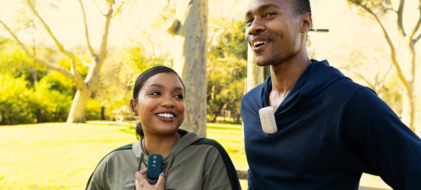 Two people running outdoors in a sunny park, one of them holding a smartphone and both wearing earpieces, smiling and talking while enjoying a jog together.