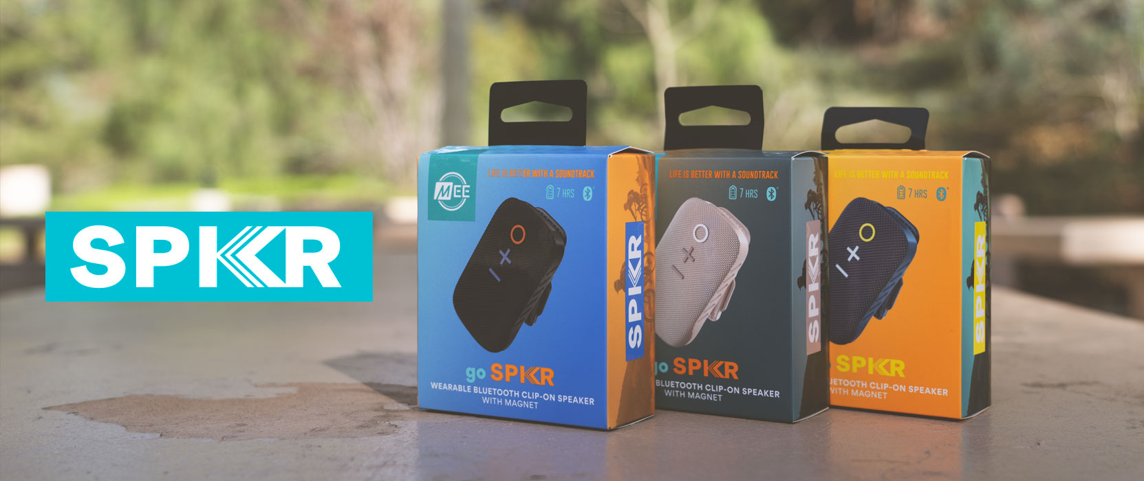 Three "spkr" brand portable speaker packages displayed on a wooden surface outdoors, with variations in design and color labeled as wood, blue, and black.