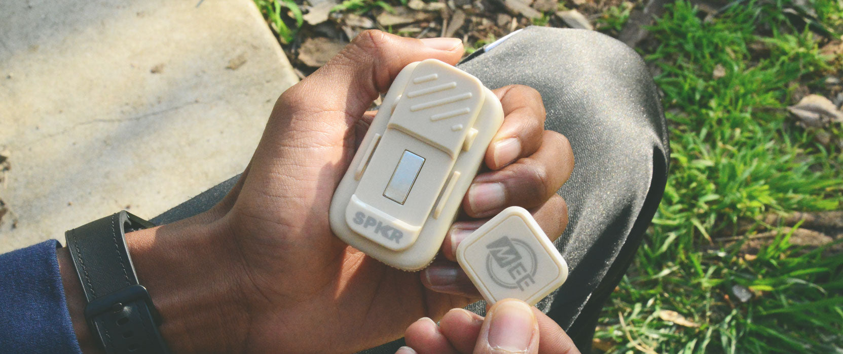 A person holding two hearing aids in their hands outdoors, with focus on the devices. one is labeled 'spk' and the other 'mic'.