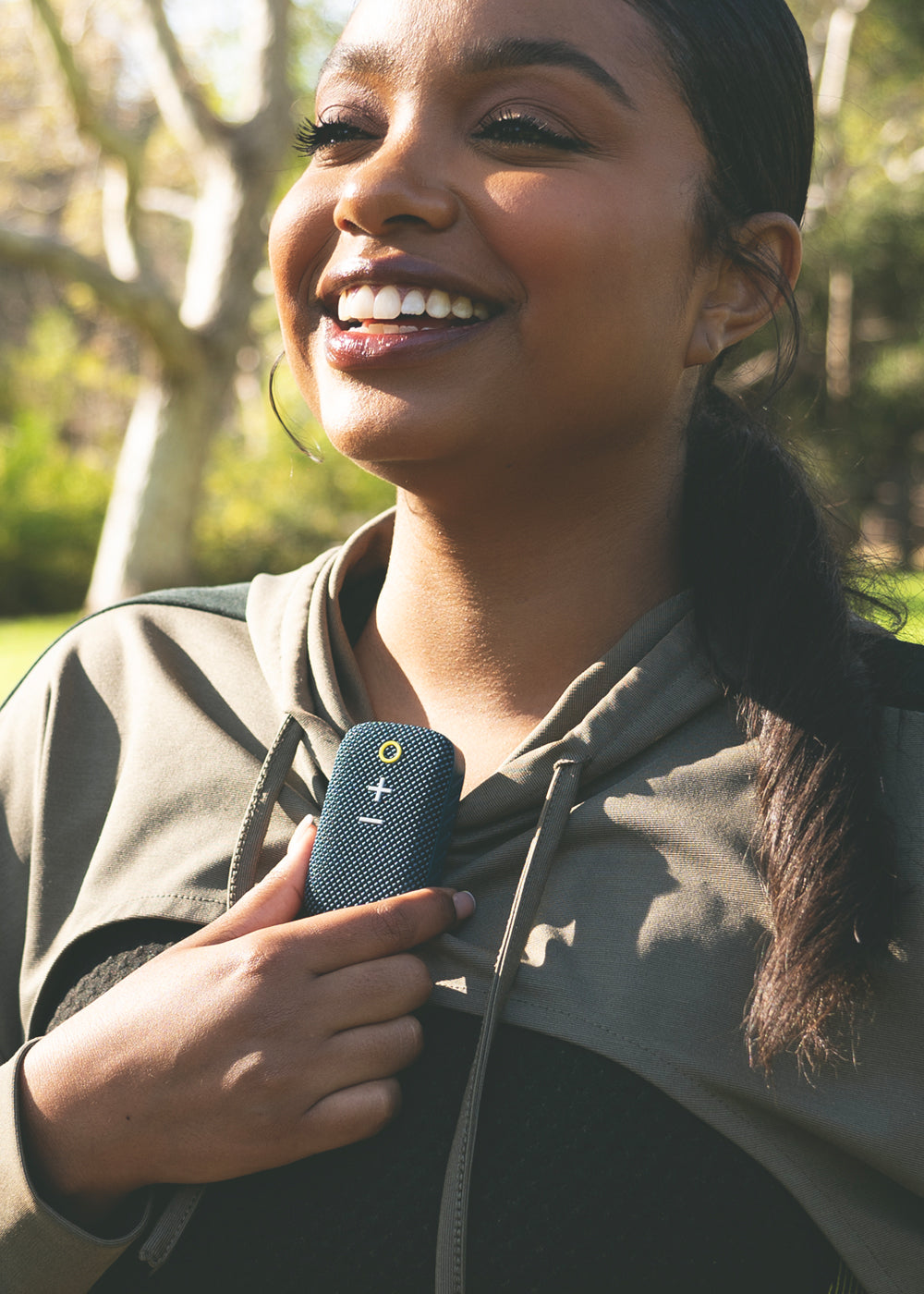 A joyful young woman holding a small blue speaker close to her chest, smiling broadly in a sunlit park. she has her hair in a ponytail and wears a sporty jacket.
