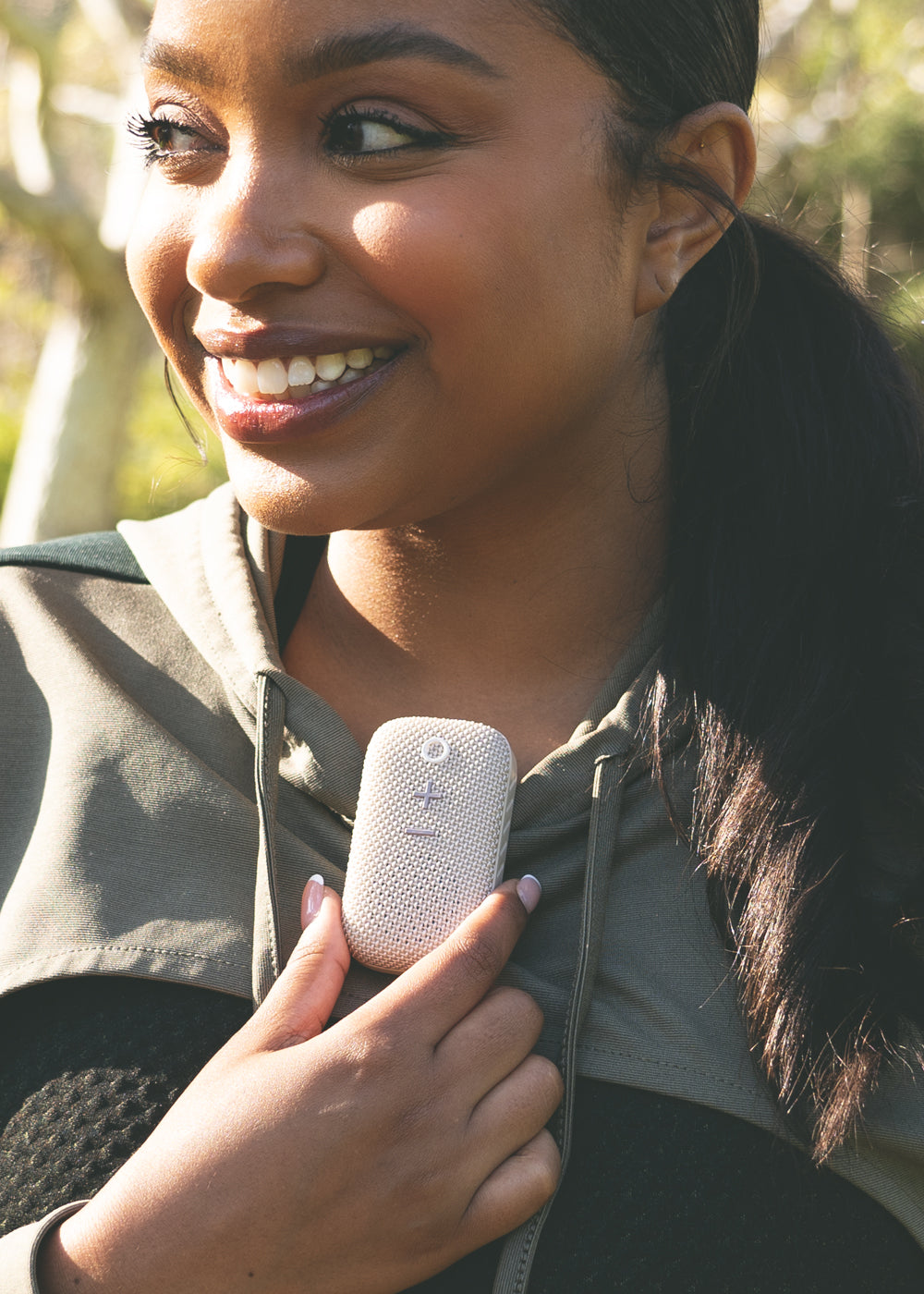 A smiling woman holding a portable speaker in her hand while standing outdoors, sunlight filtering through trees in the background.