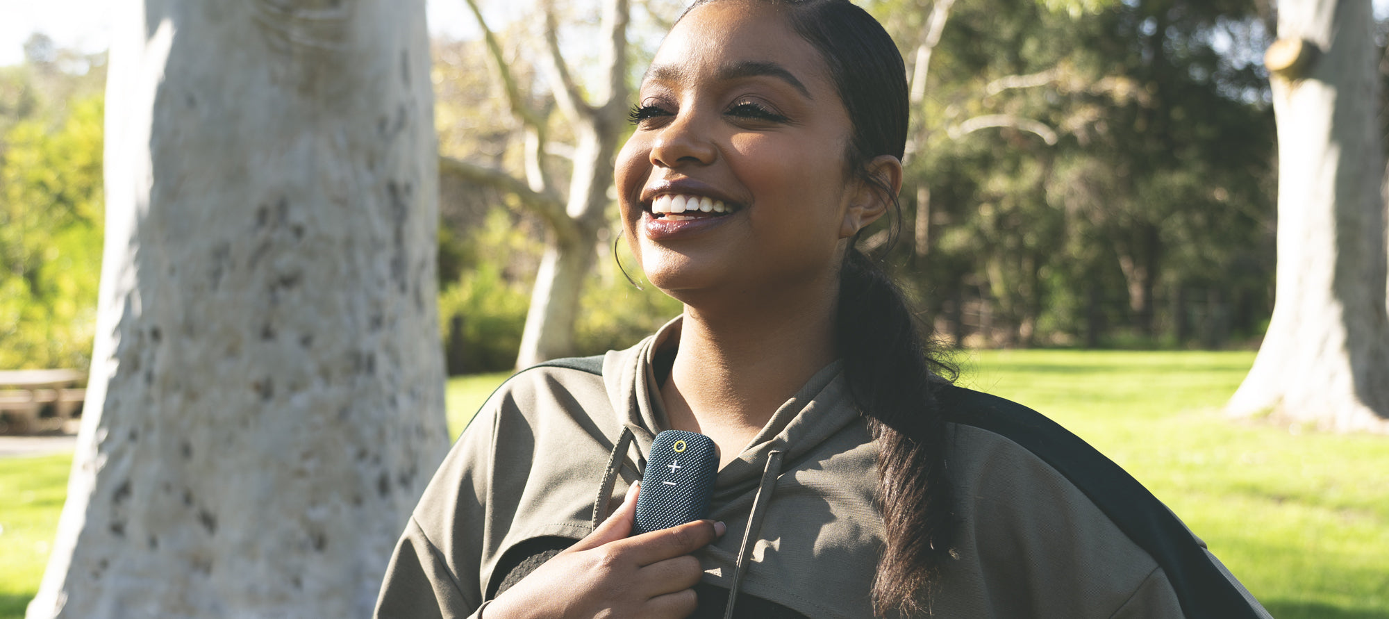 Smiling woman with a ponytail holding a smartphone, standing outdoors in a sunlit park with trees in the background.