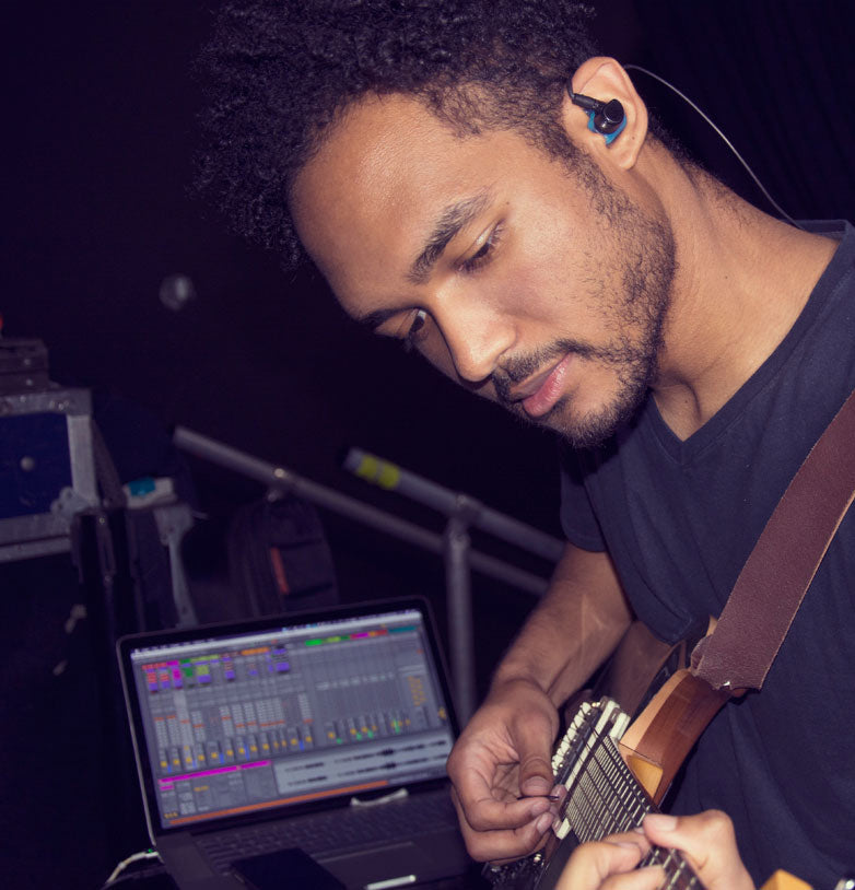 A young man with curly hair, wearing in-ear headphones, intently plays an electric guitar near a laptop displaying music production software.