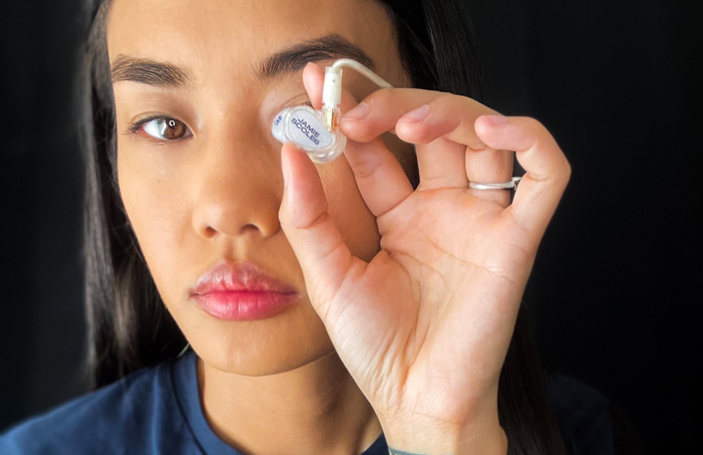 A young woman holding a small, labeled ampoule close to her eye, focusing intently on it. she has a neutral expression and a black background enhances the clarity of the image.