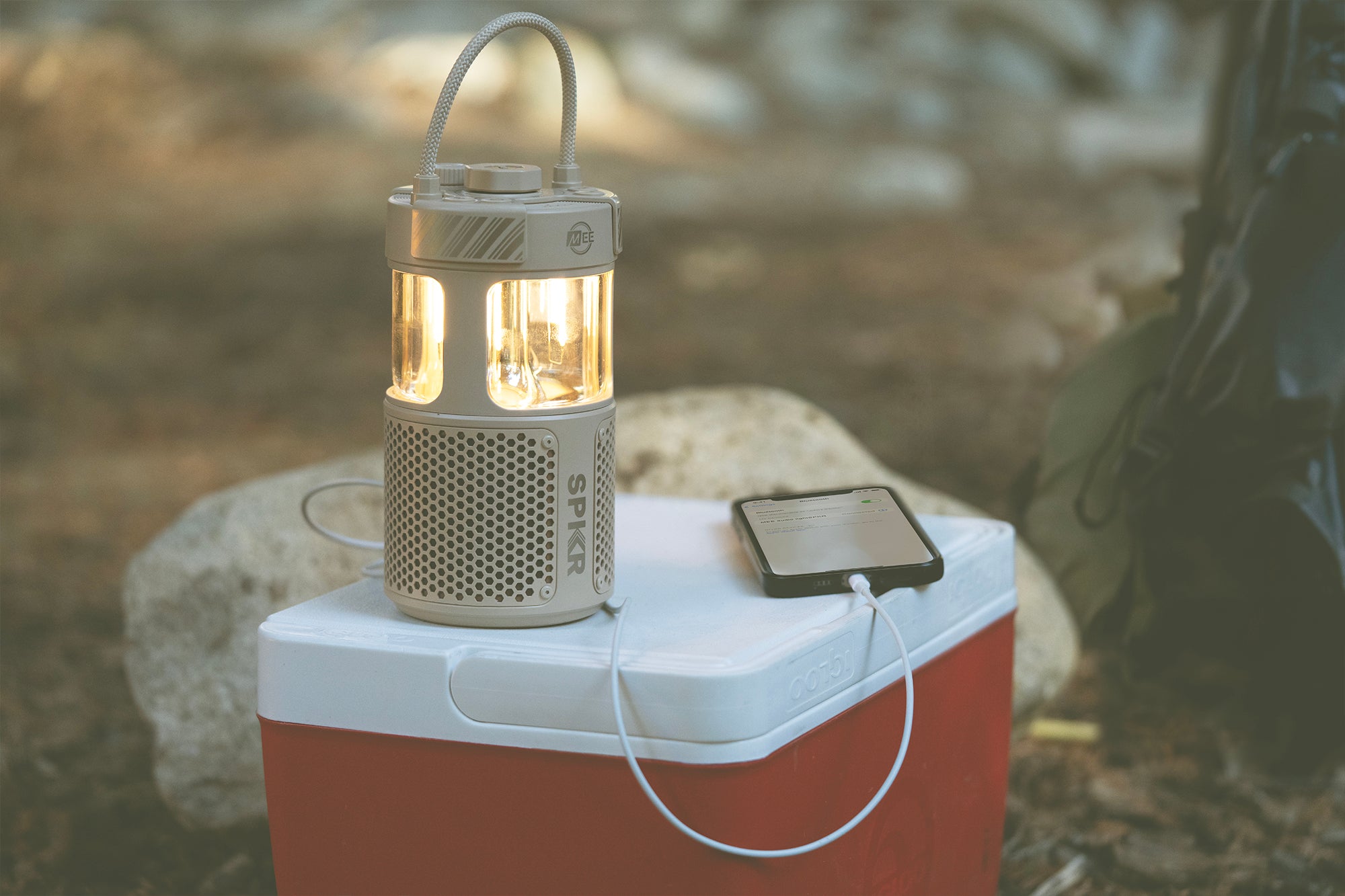 A portable lantern on a red and white cooler in a forest setting, connected by a cable to a smartphone, suggesting the lantern is charging the phone.