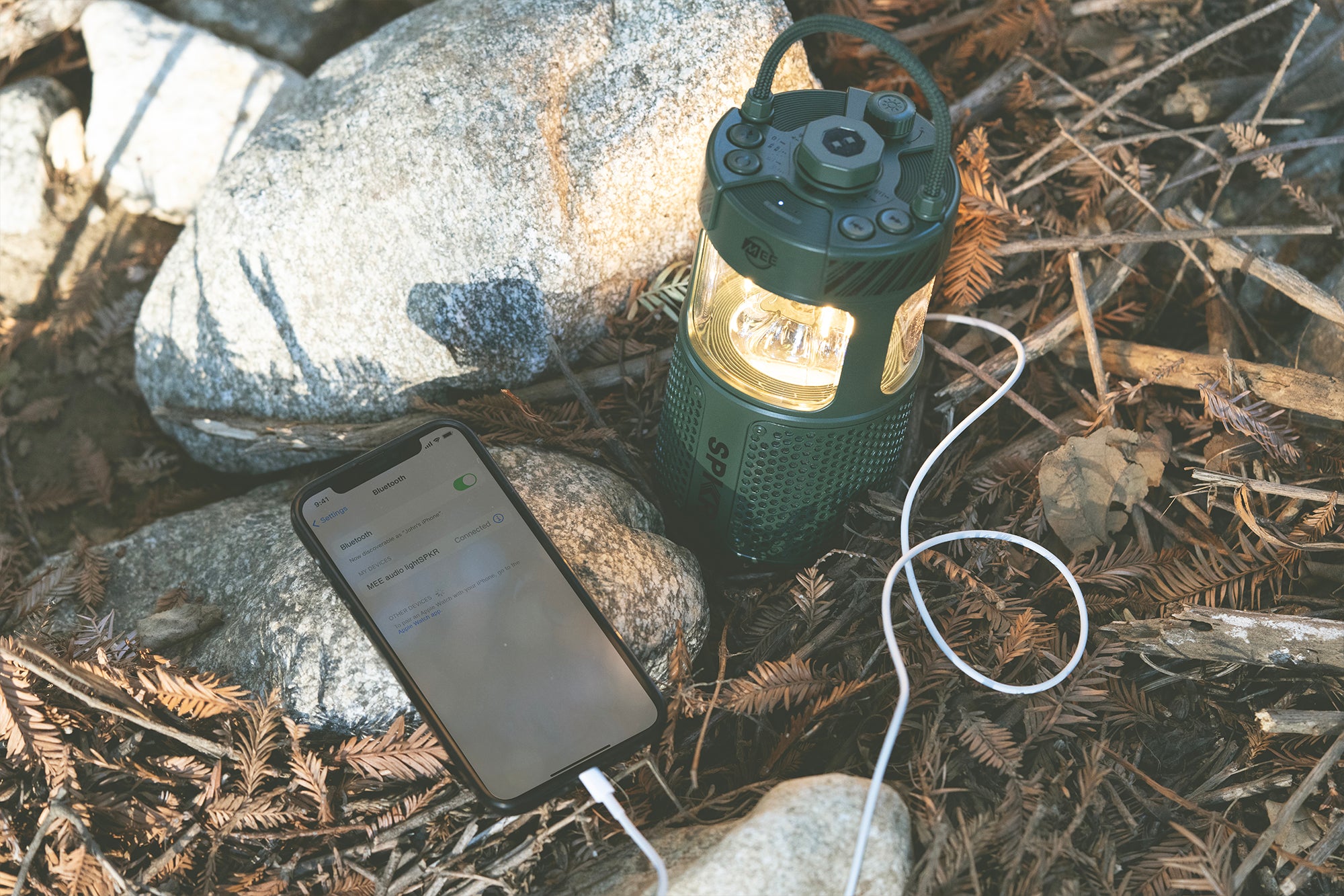 A smartphone connected via usb to a portable, green lantern on a forest floor among stones and fallen leaves, displaying an active charging screen.