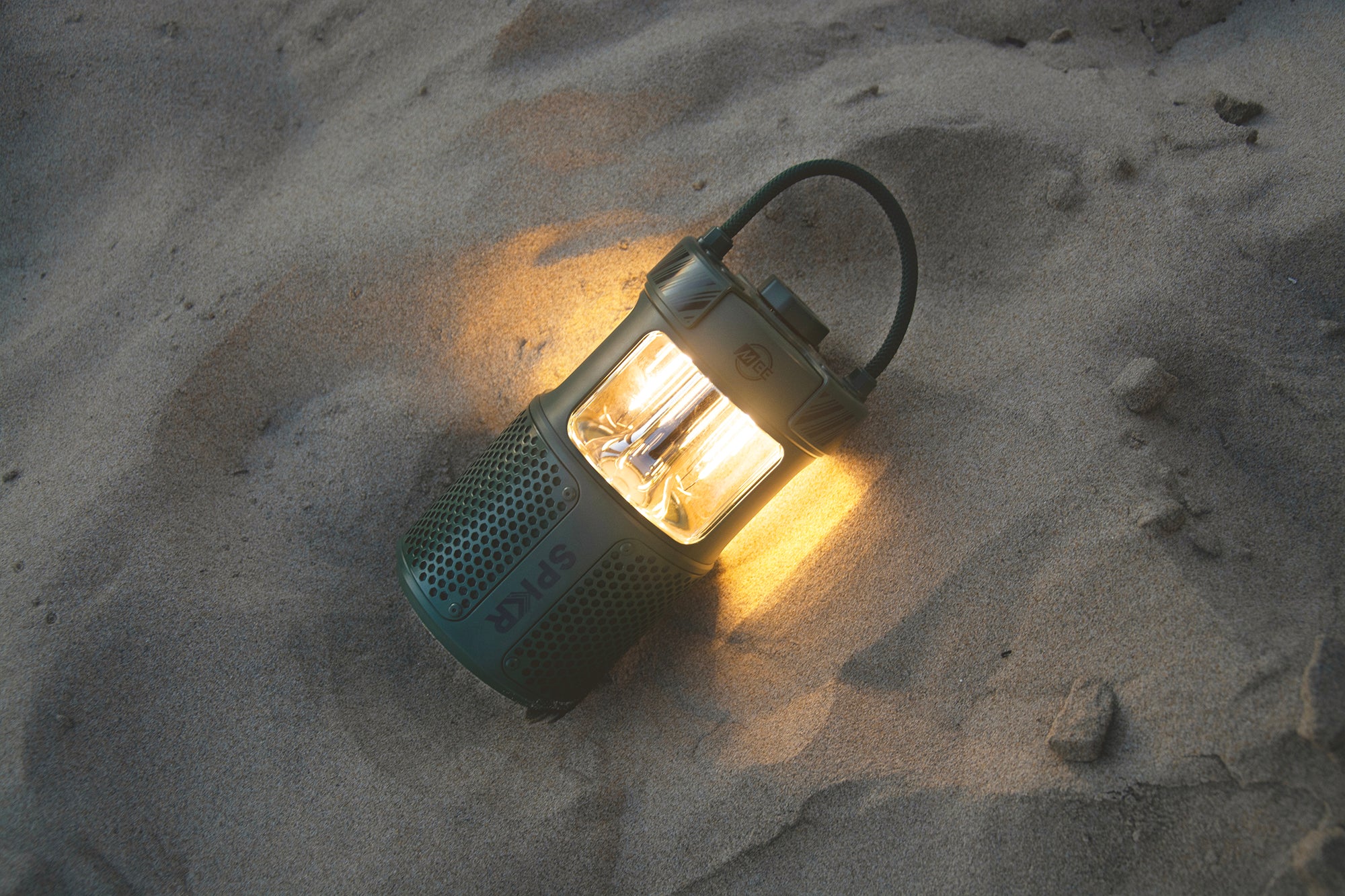 A glowing lantern sits on sandy beach soil, casting a warm light during dusk, highlighting the intricate design and the brand label on its side.