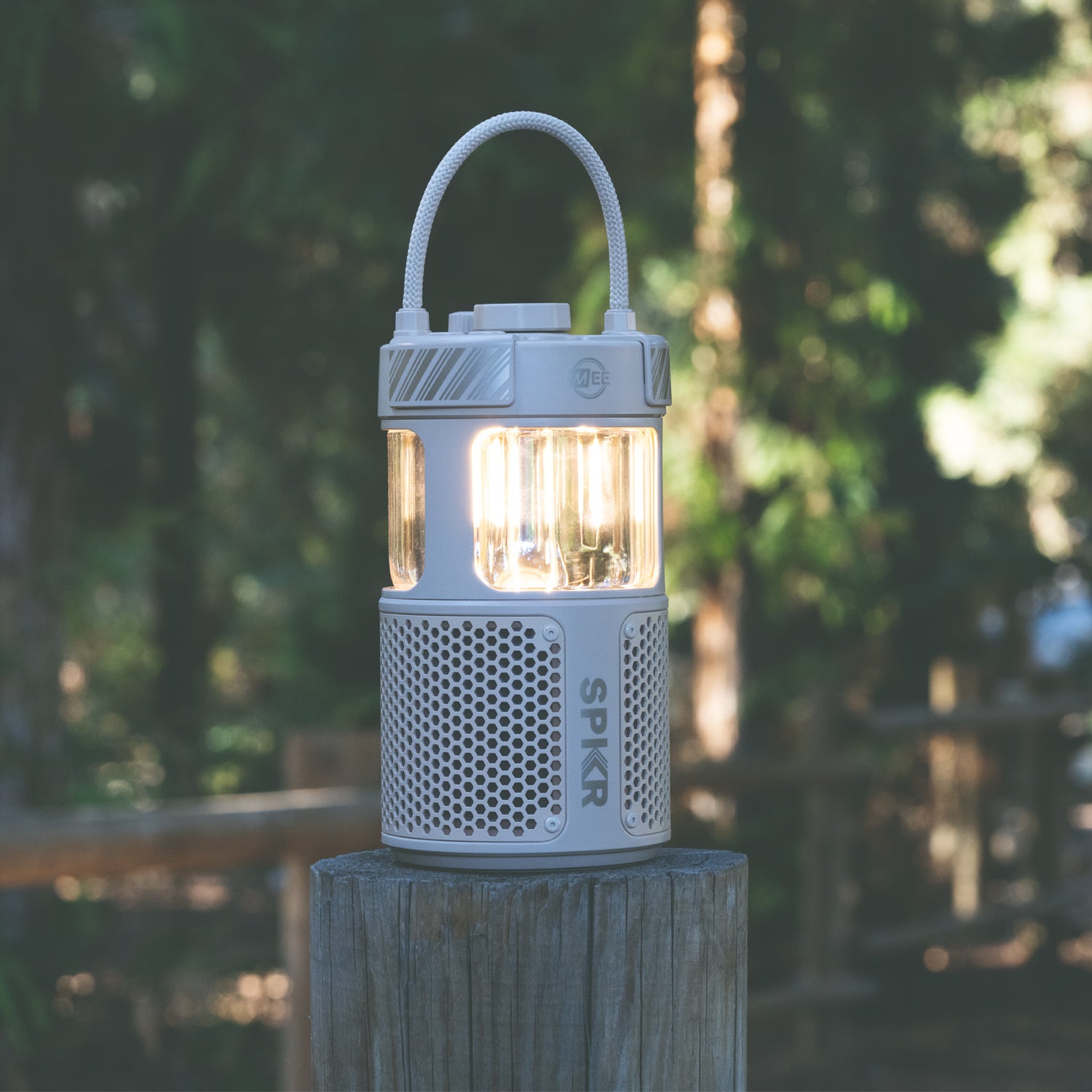 A portable led lantern sits atop a wooden post in a forest, emitting a warm glow with trees softly blurred in the background. the lantern is white with a gray handle and marked with the logo "spkr.