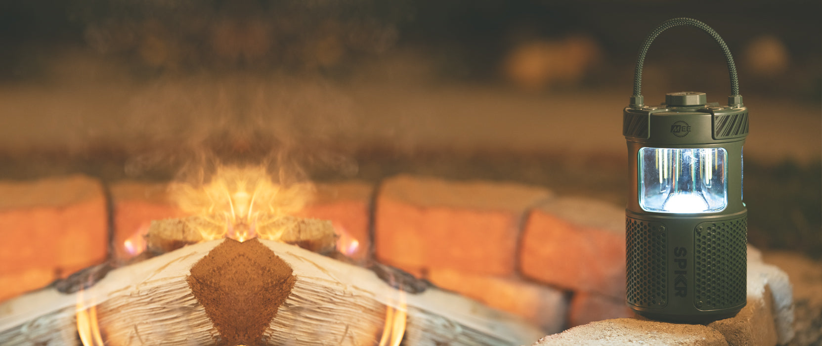 A green led camping lantern stands illuminated near a log on fire surrounded by sand-colored bricks, emitting a warm glow with smoke drifting upwards in a cozy outdoor setting.