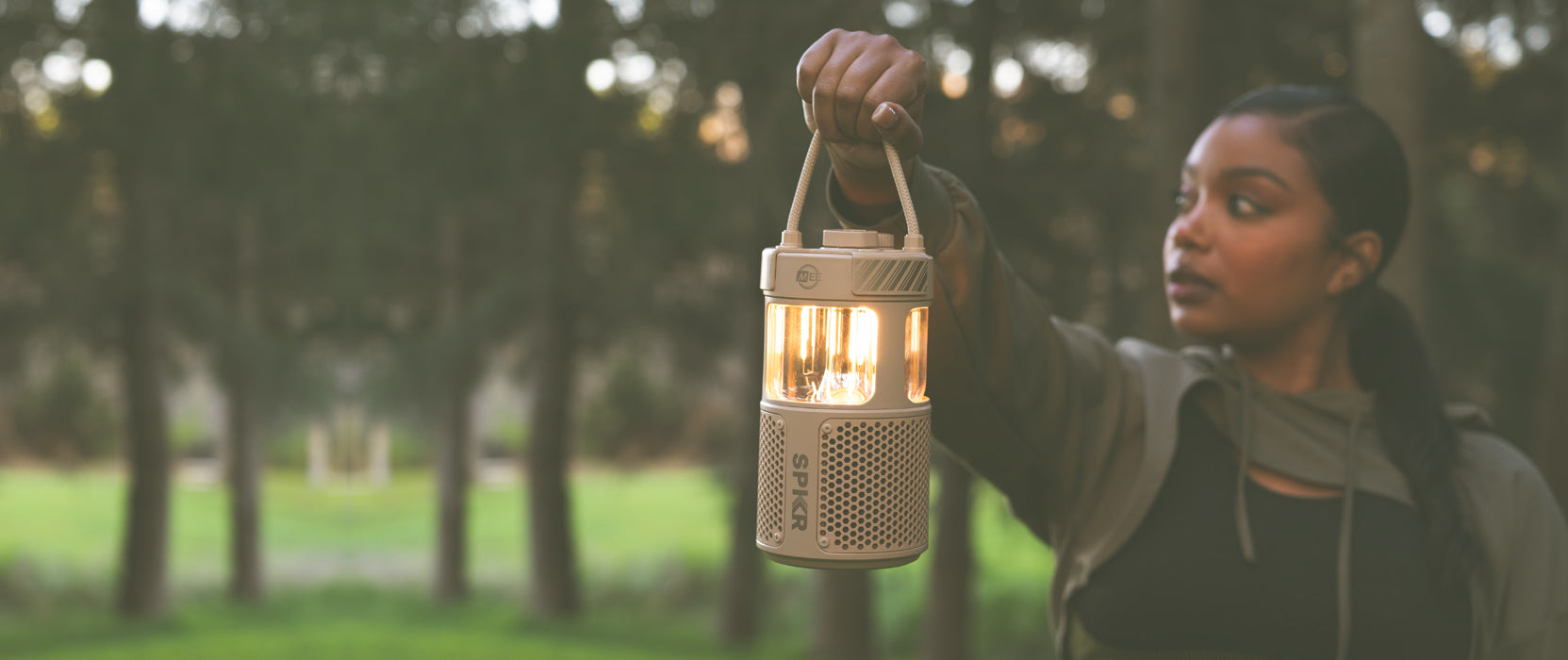 A woman holding a glowing lantern in a forest at dusk, focusing on the light with a serene expression, surrounded by trees in soft focus.
