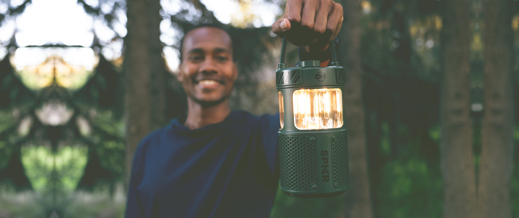 A man smiling in a forest, holding a lit lantern closely towards the camera. the background is softly blurred with green trees and sunlight filtering through.