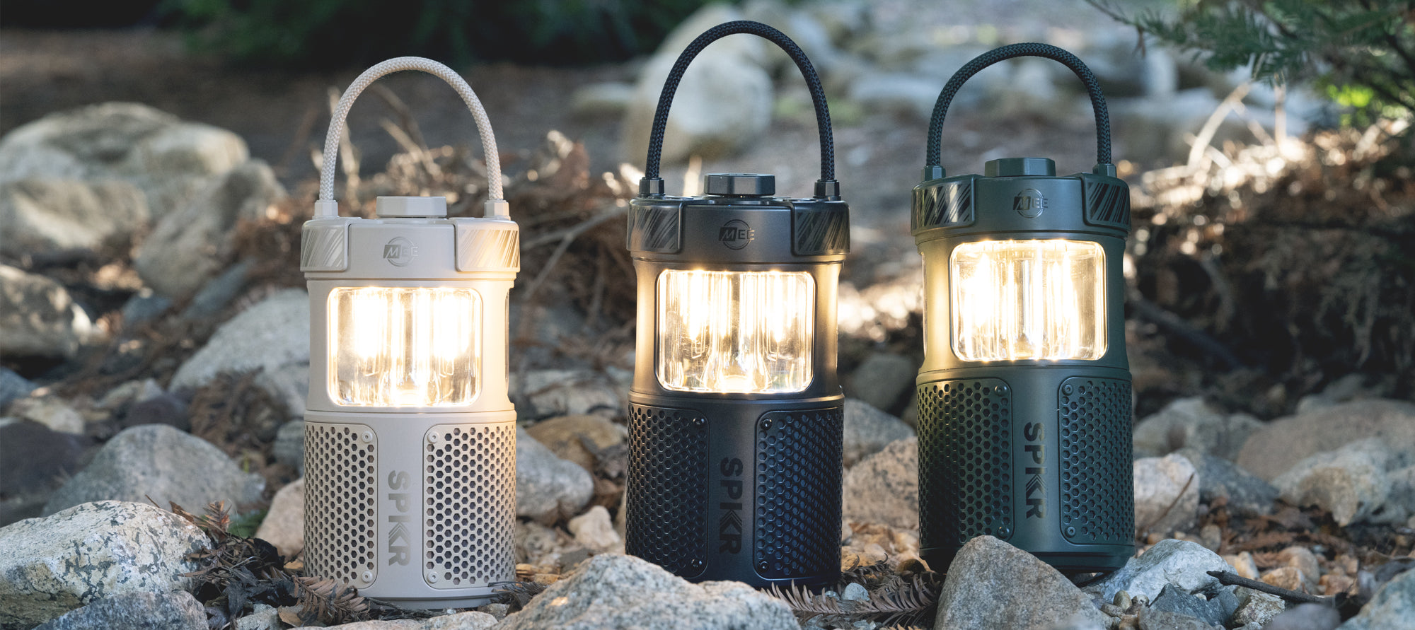 Three portable led lanterns, in cream, black, and green, illuminated and standing on a rocky ground with sparse vegetation in the background.