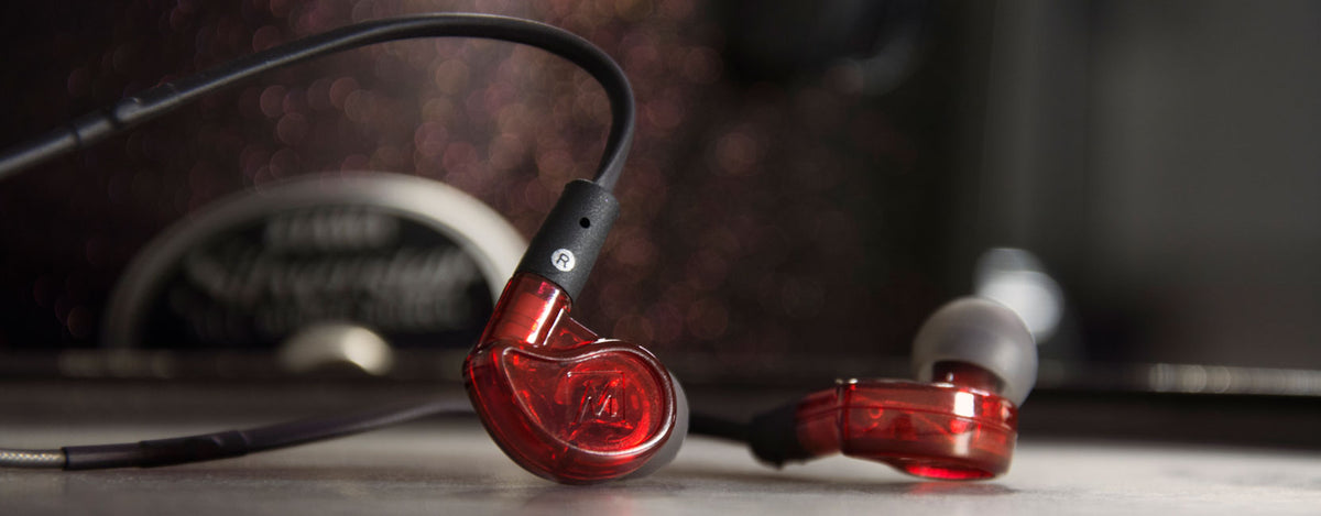 Close-up of red M6 PRO in-ear monitors with a black audio cable, resting on a reflective surface against a blurred background with dark tones.