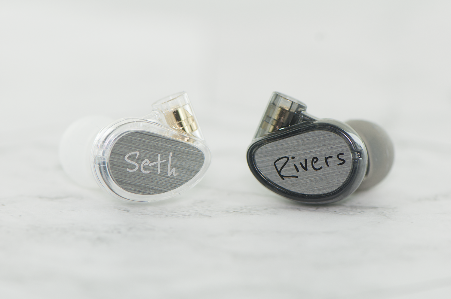 Two custom earphones with names "seth" and "rivers" inscribed on metallic surfaces, lying against a white marble background.