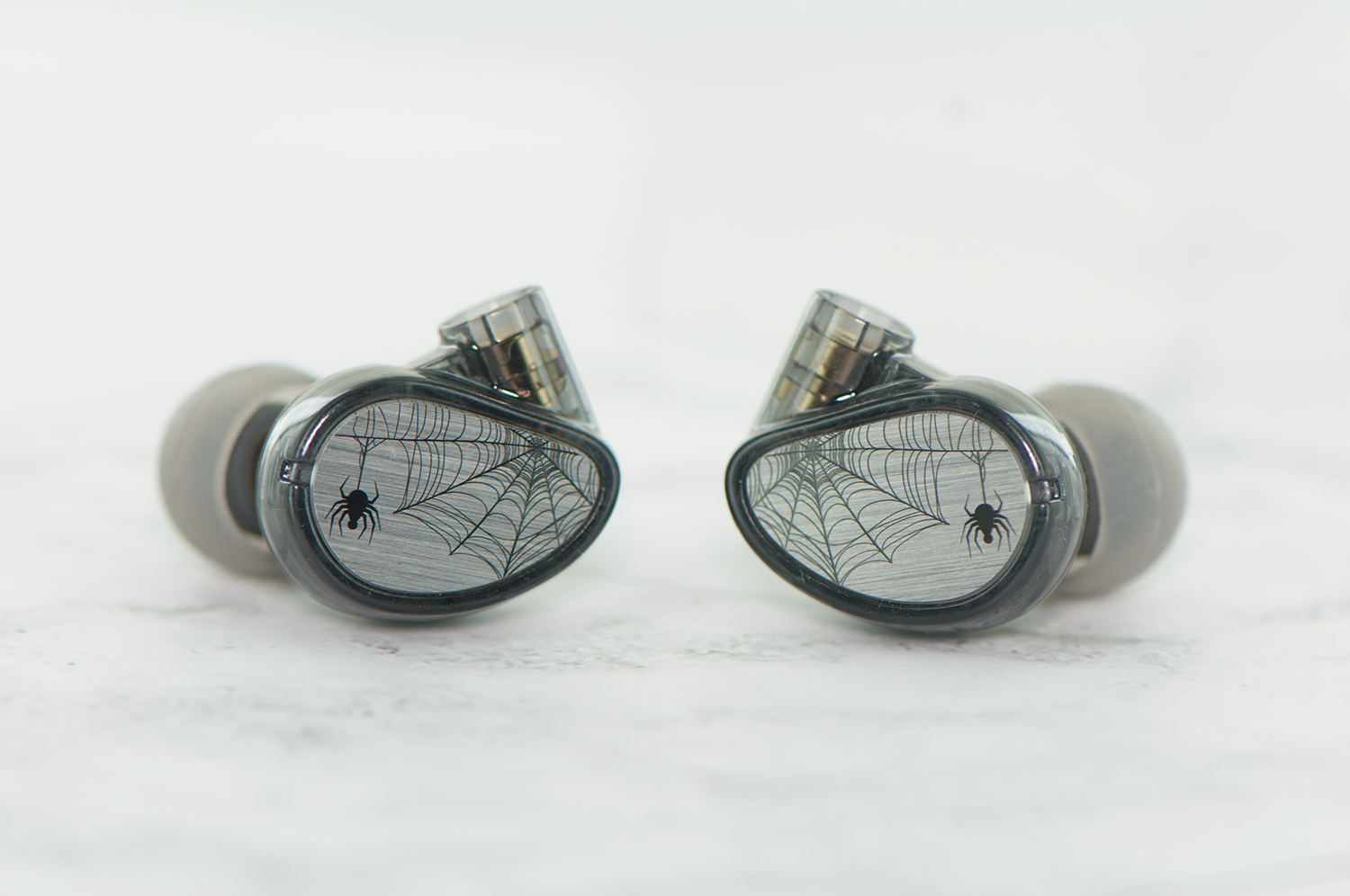 Two high-end earphones with a spider and web design on a reflective metallic surface, set against a soft white background.
