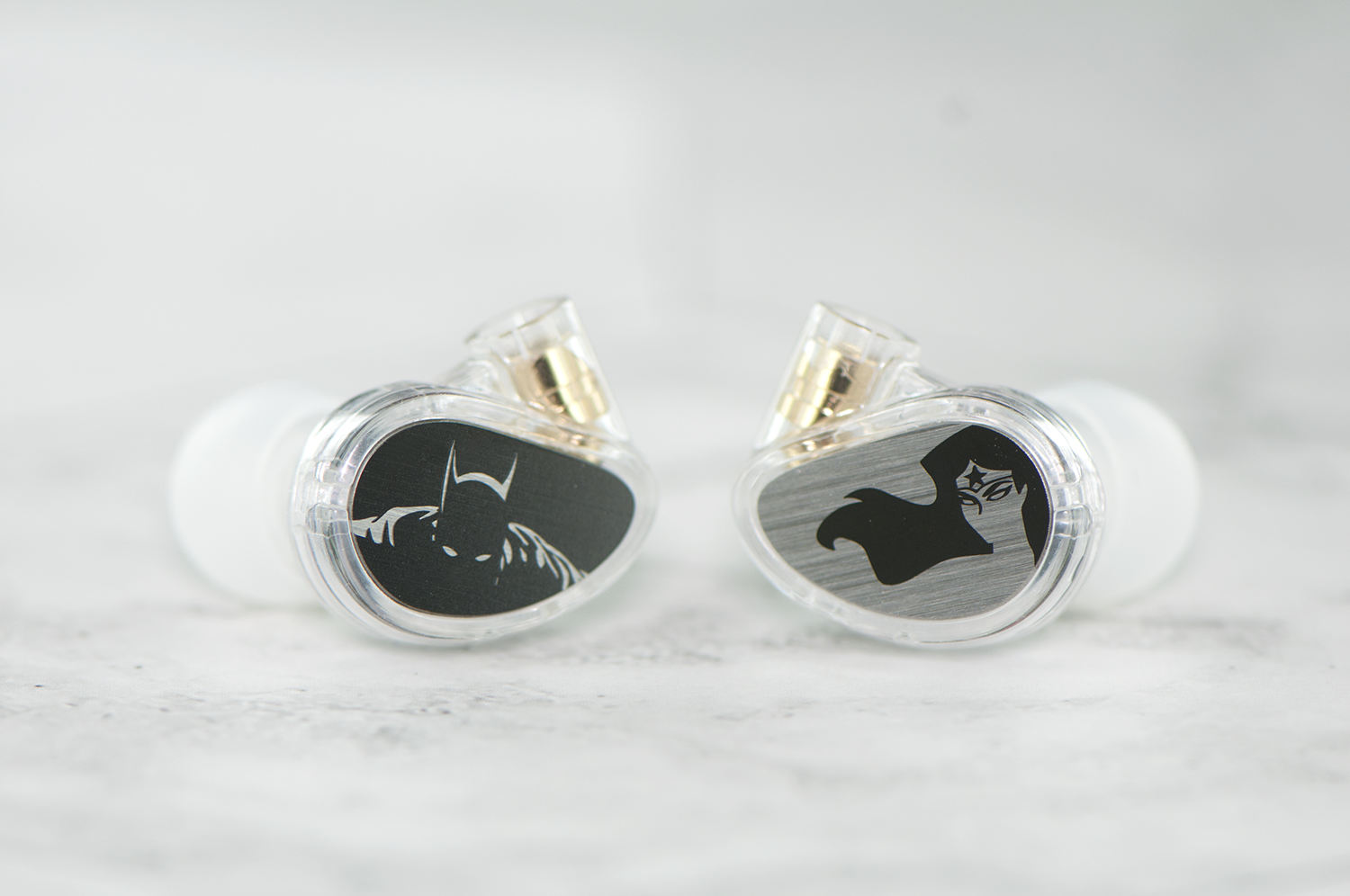Two in-ear monitors with clear silicone tips and transparent casings, showcasing custom black and silver superhero designs on the back plates, resting on a marble surface.