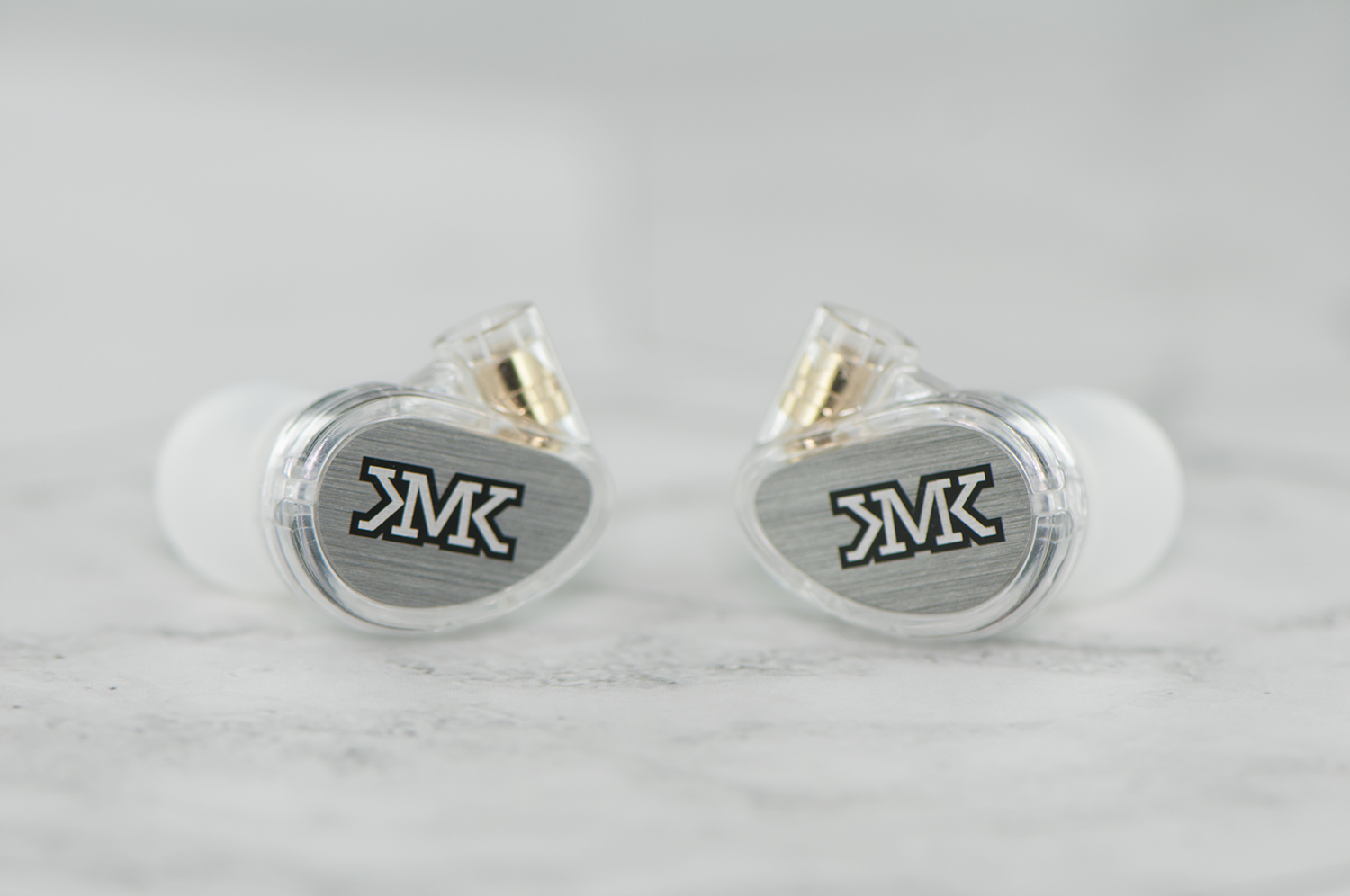 Two transparent in-ear monitors with a "jmk" logo, resting on a gray marble surface, showcasing a metallic and clear design with gold-tone accents.