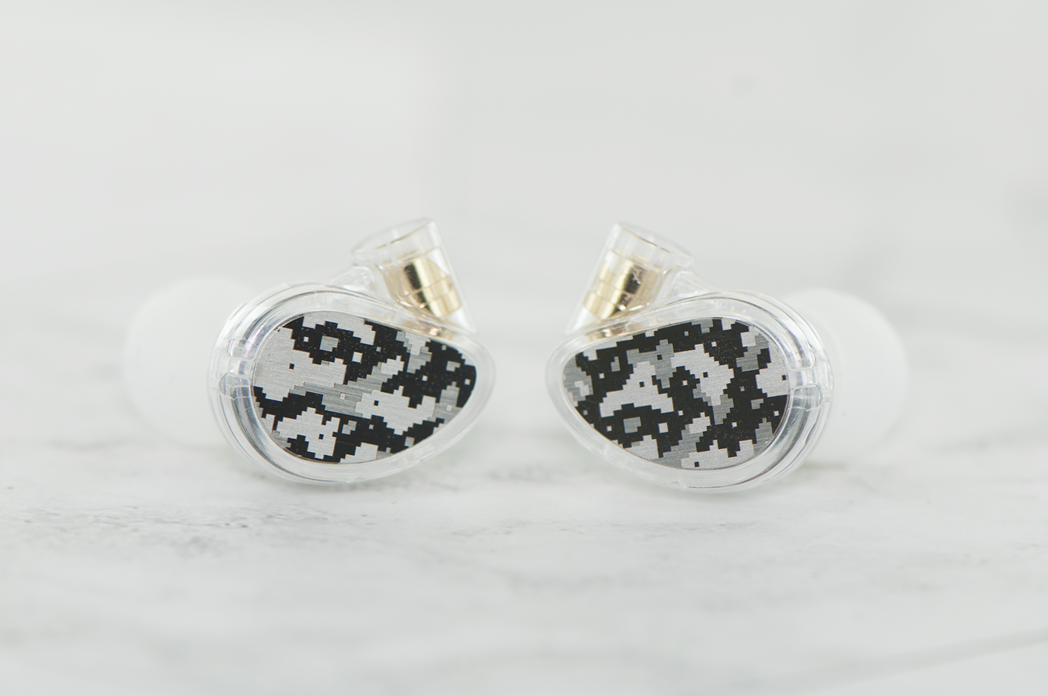 A pair of transparent in-ear monitors with a unique pixelated design on the faceplates, displayed against a light gray background.