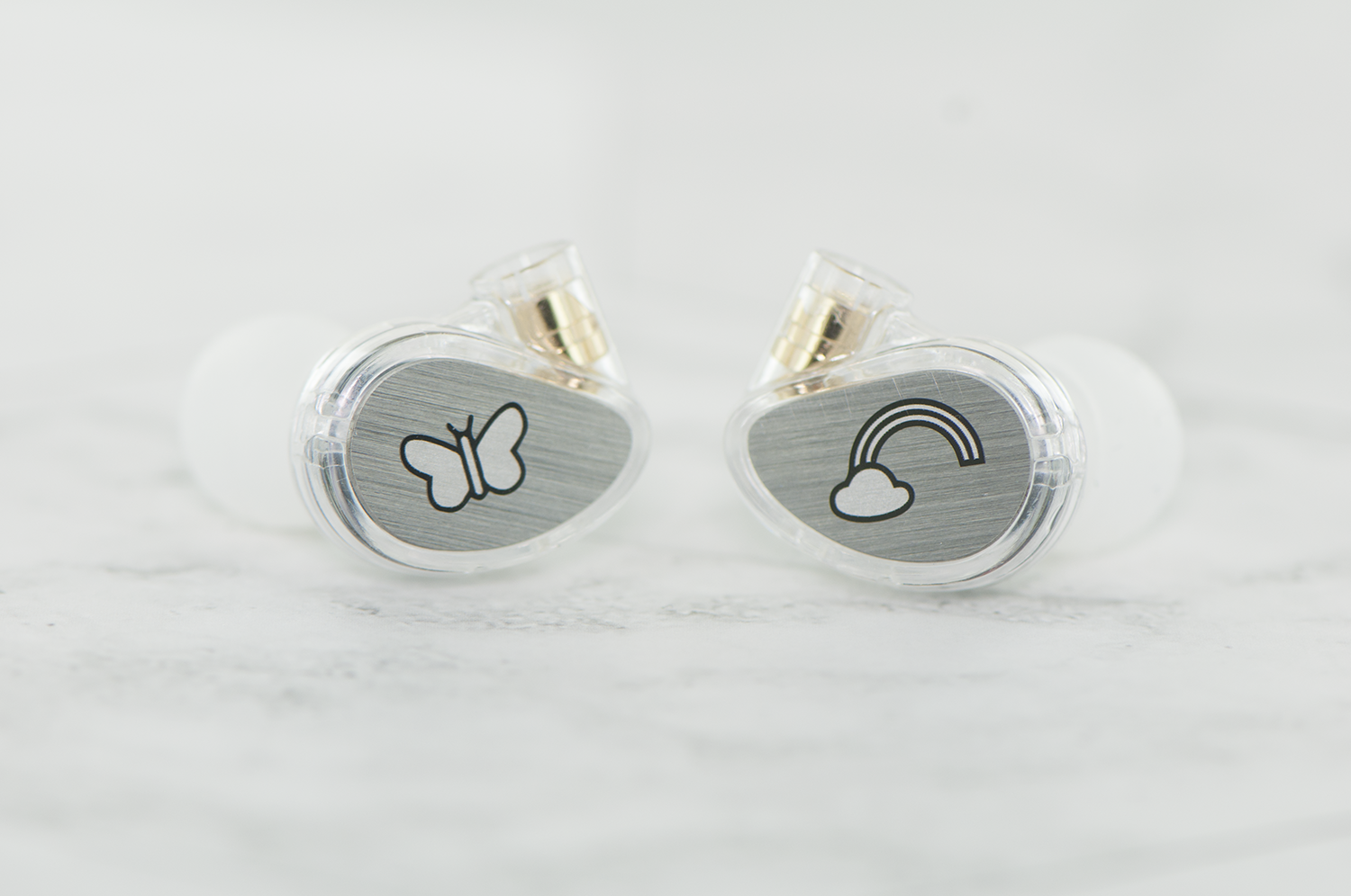 Two in-ear monitors with transparent casing, showcasing internal components and unique designs—a butterfly on one, and a cloud with a rainbow on the other, on a white surface.