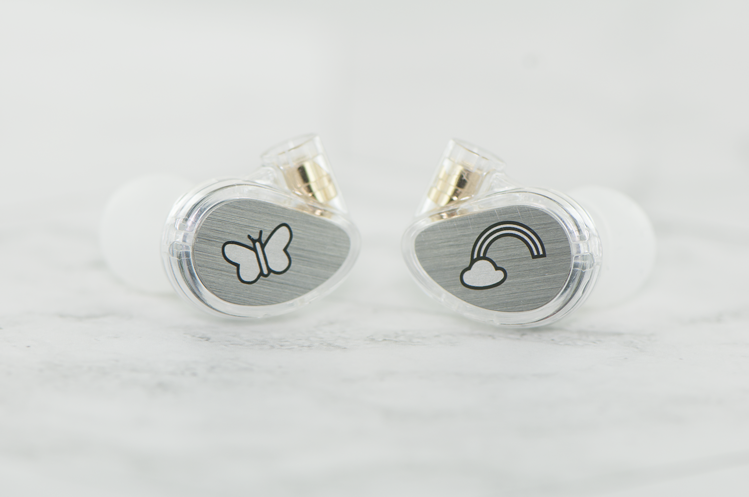 Two in-ear monitors with transparent casing, showcasing internal components and unique designsâ€”a butterfly on one, and a cloud with a rainbow on the other, on a white surface.