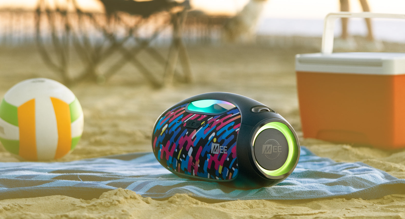 A colorful portable speaker on a sandy beach with a volleyball and a cooler in the background, capturing a relaxed beach day setting.