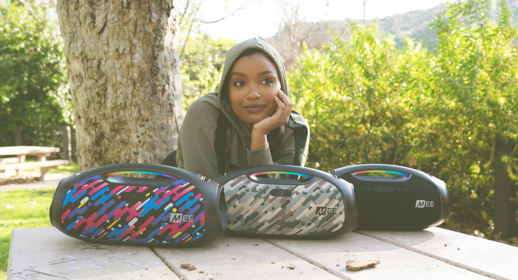 A young woman in a hijab smiling by a wooden table, with three colorful portable speakers in front of her, outdoors with trees and sunlight in the background.