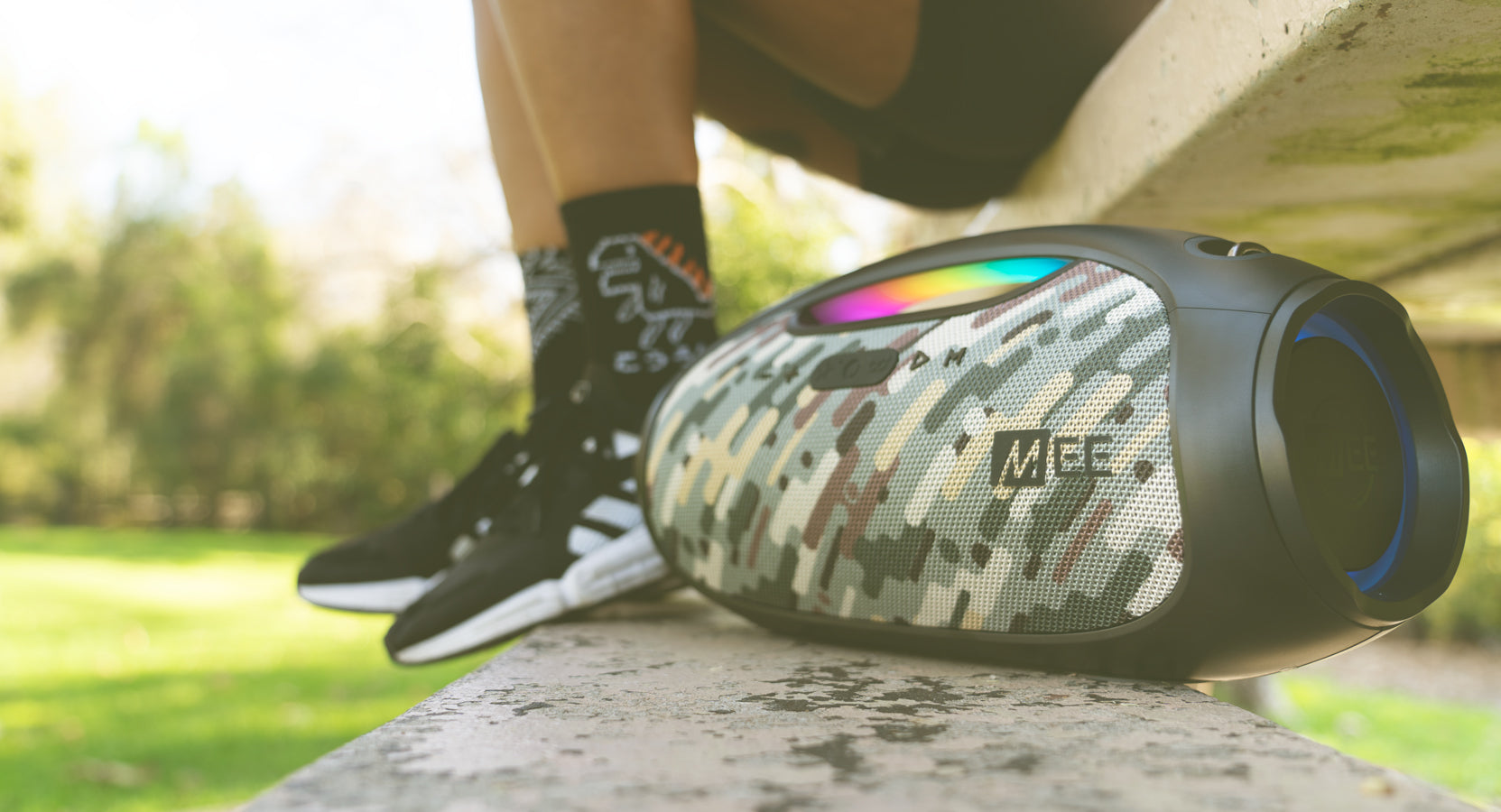 A portable camouflage speaker sits on a concrete ledge outdoors next to a person wearing sneakers and patterned socks. the speaker is lit up with colorful lights.