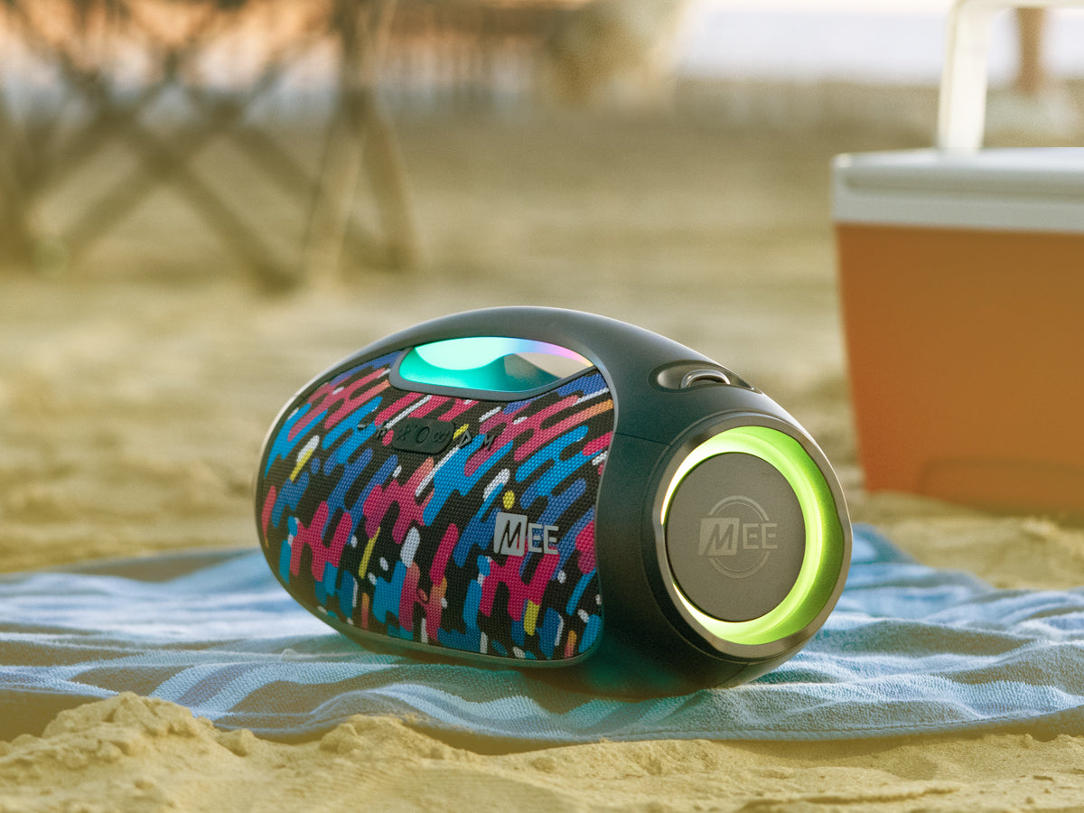 A colorful portable speaker on a sandy beach with a cooler in the background, emitting a green glow under warm sunlight.