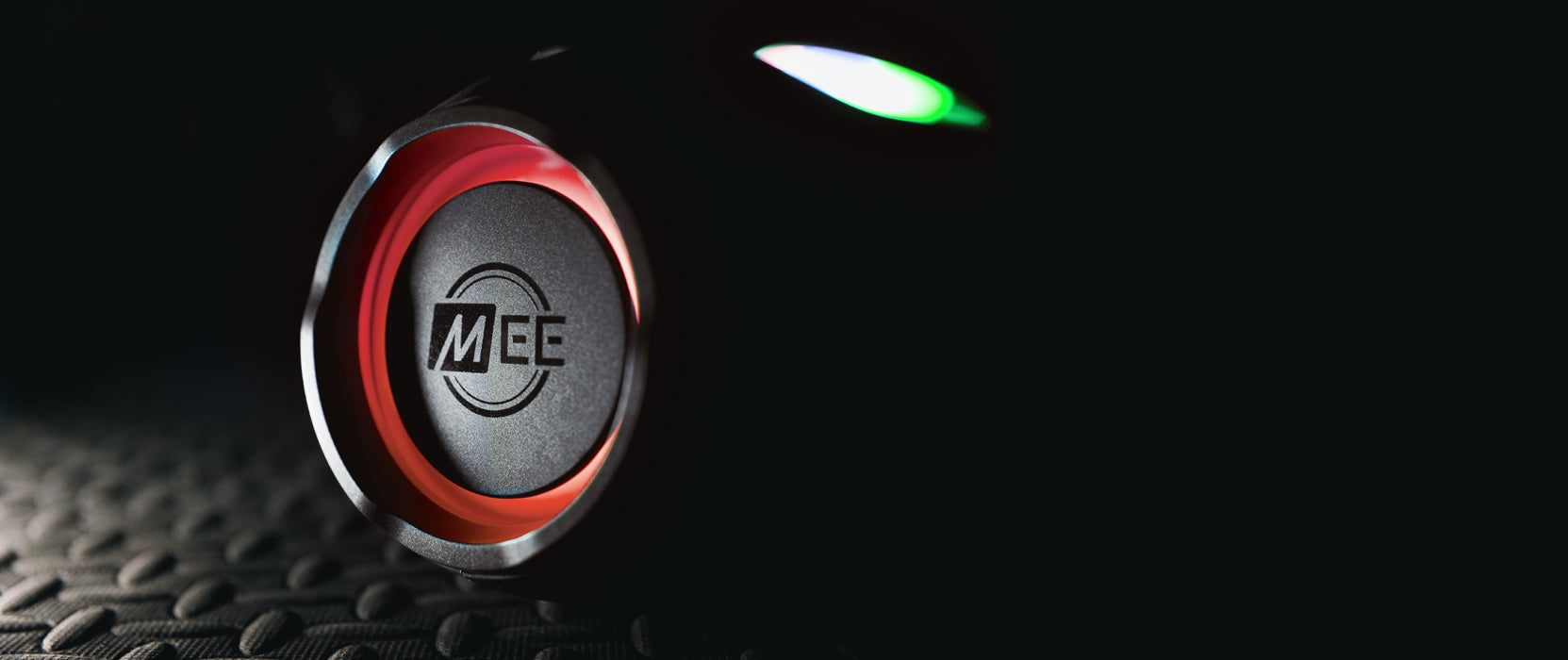 Close-up view of a circular, red push button with "me" logo, illuminated by a soft green light, set against a textured, dark background.