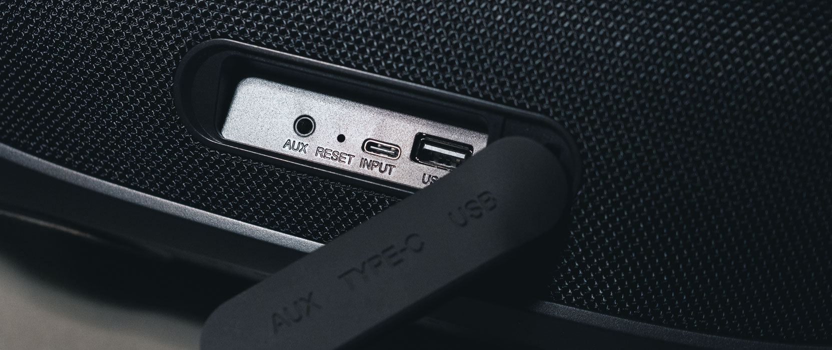 Close-up of a speaker's connectivity panel featuring an auxiliary input, usb port, and a reset button, all under a labeled rubber cover.