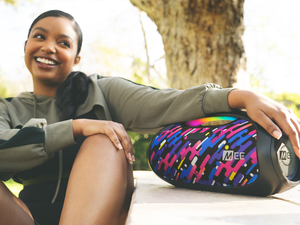 A smiling woman sitting outdoors next to a colorful portable speaker, enjoying a sunny day in a park.