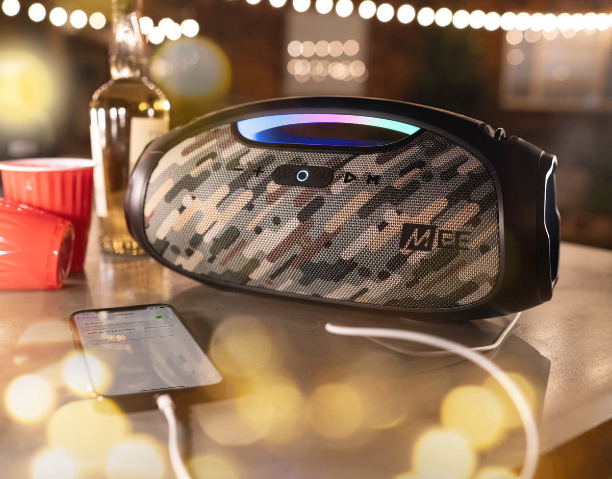 A portable camouflage-patterned speaker connected to a smartphone on a wooden table, with a blurred background of glowing lights and a bottle.