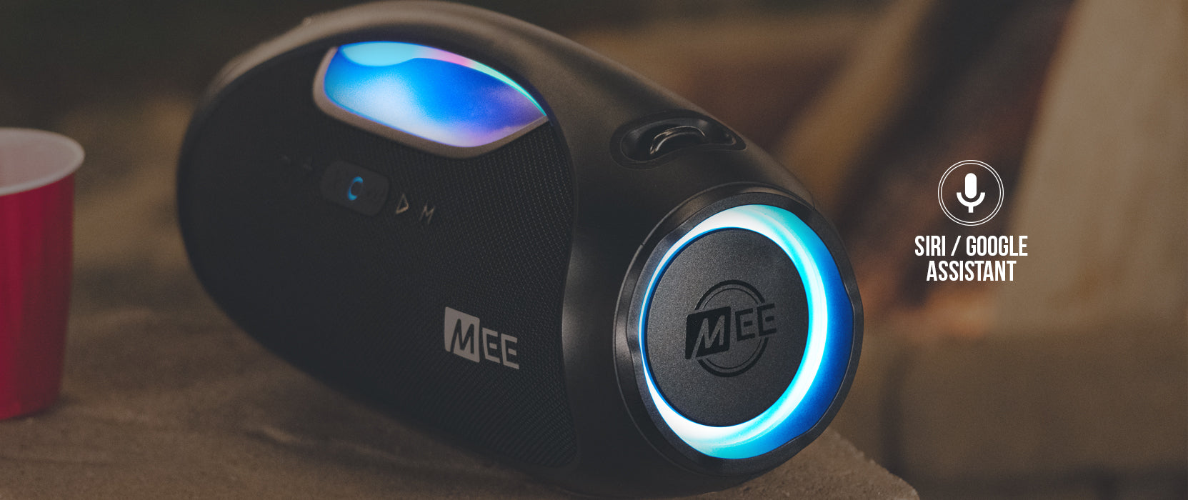 Portable wireless speaker with blue led lights, featuring voice assistant compatibility icons for siri and google assistant, displayed on a wooden table.