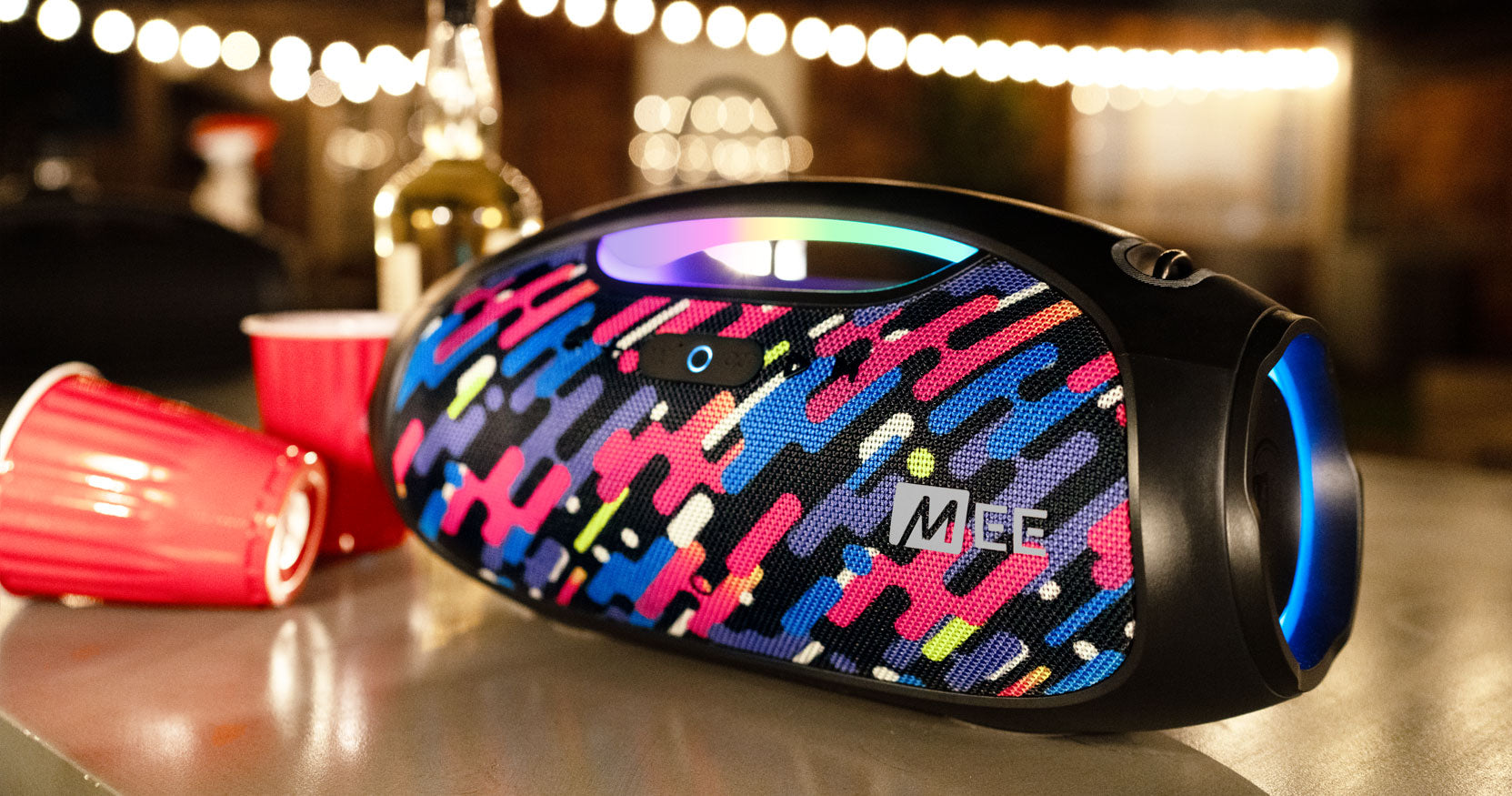 A colorful portable speaker with a vibrant abstract pattern on a table, surrounded by party cups and a wine bottle, illuminated by string lights in the background.