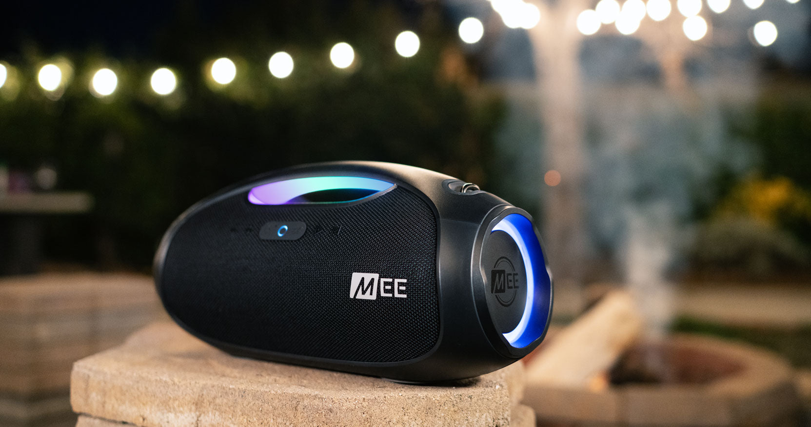 Portable bluetooth speaker with vibrant blue illumination, placed on an outdoor stone ledge, illuminated by background string lights at dusk.