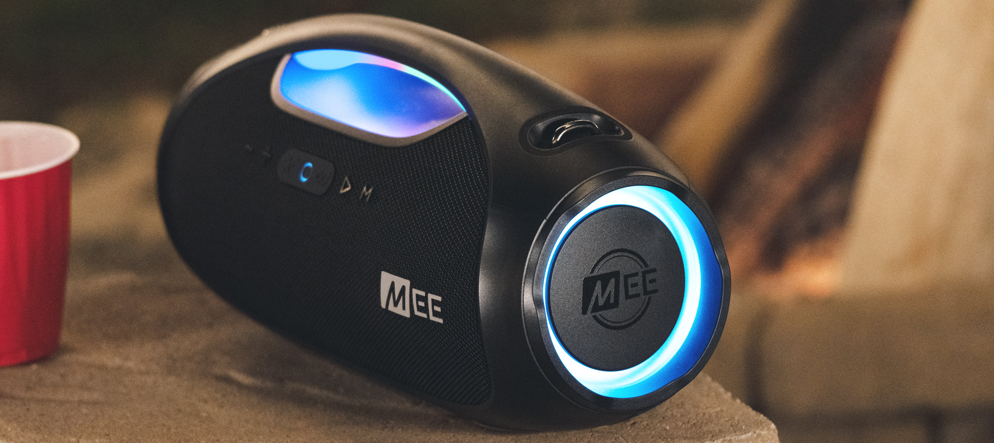 A portable bluetooth speaker with blue led lights glowing around the speaker grills, placed on a wooden surface near a red cup.
