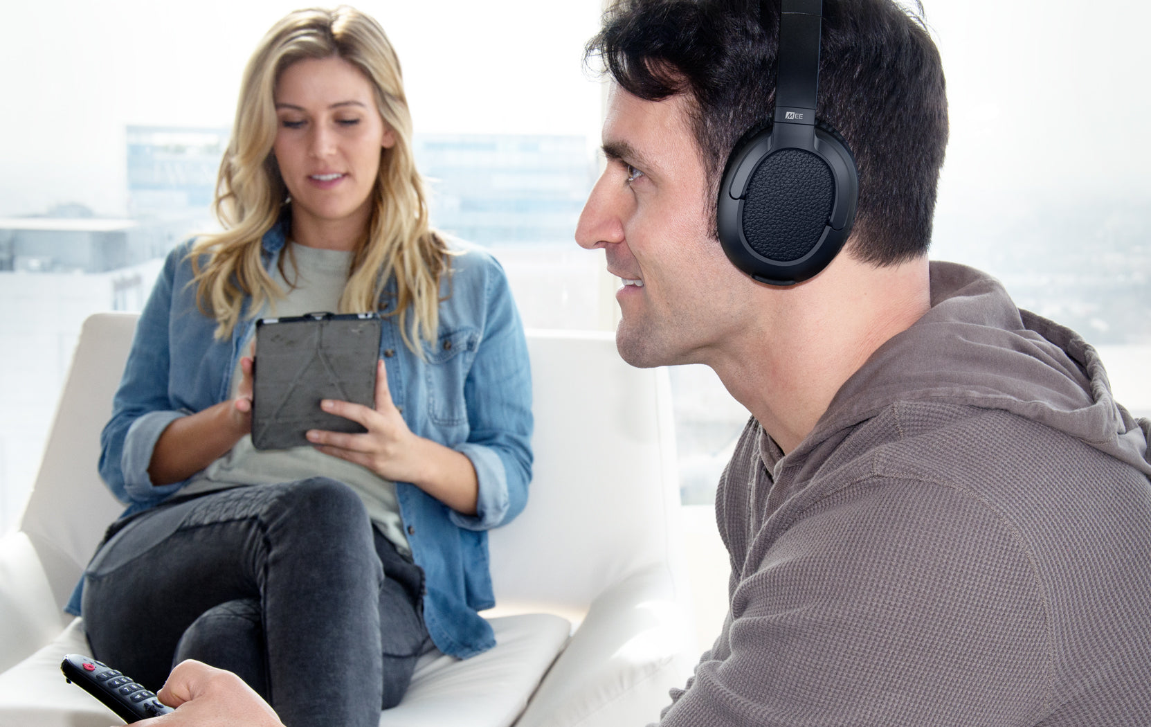 A man wearing headphones sits next to a woman holding a tablet in a bright room with a white couch, both appearing engaged in separate activities.