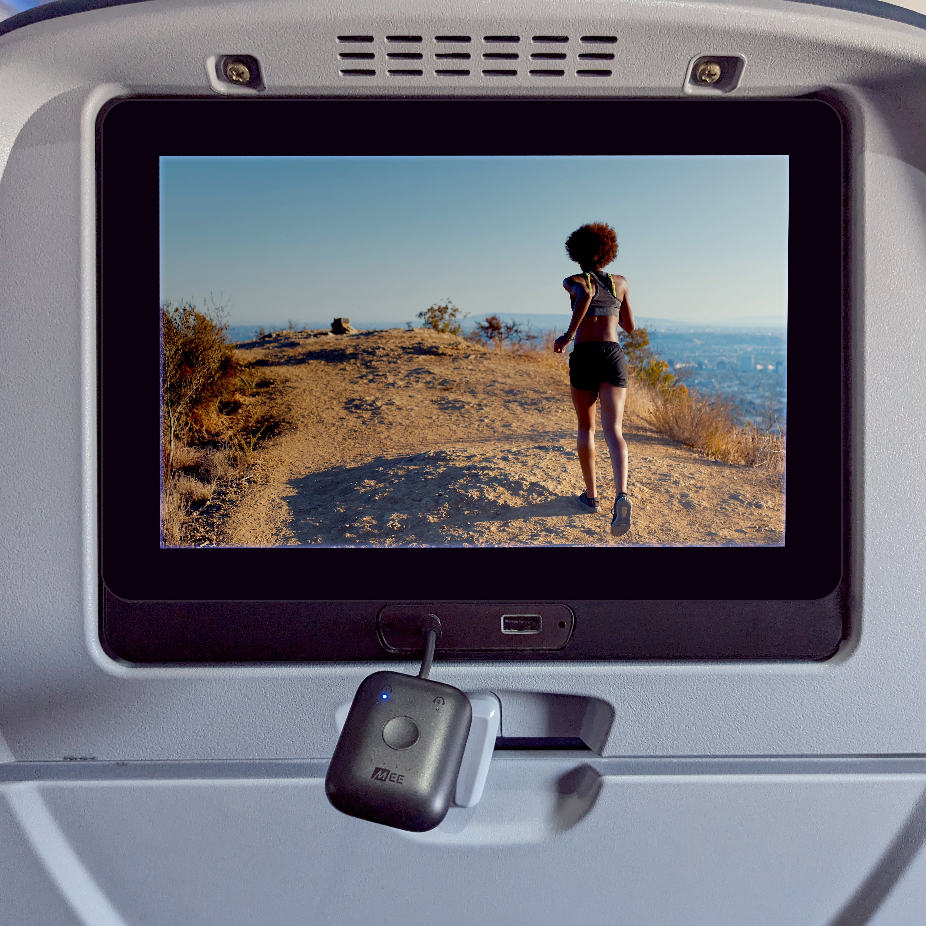 A car entertainment system screen displays an image of a young woman jogging on a mountain trail, overlooking a cityscape under a clear sky. an aux cable hangs in front of the screen.