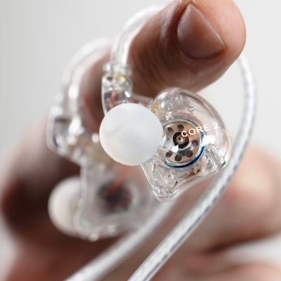 A close-up of a hand holding the MEE audio CORE 2 dual driver hybrid transparent in-ear monitor earpiece with a white silicone tip and visible internal components, including the text 'CORE 2'