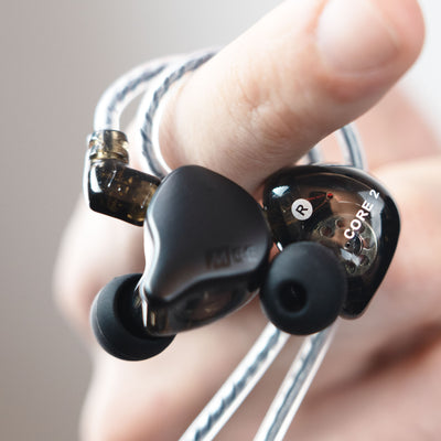 A close-up of a hand holding a pair of MEE audio CORE 2 dual hybrid driver in-ear monitors, showing the transparent dark gray earpieces with black silicone tips and braided cable. The earpiece on the right has 'CORE 2' and 'R' printed on it.
