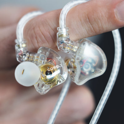 A close-up of a person holding MEE audio CORE 3 triple hybrid driver transparent in-ear monitors with visible internal components and white ear tips. The word CORE 3 is printed on one earpiece.