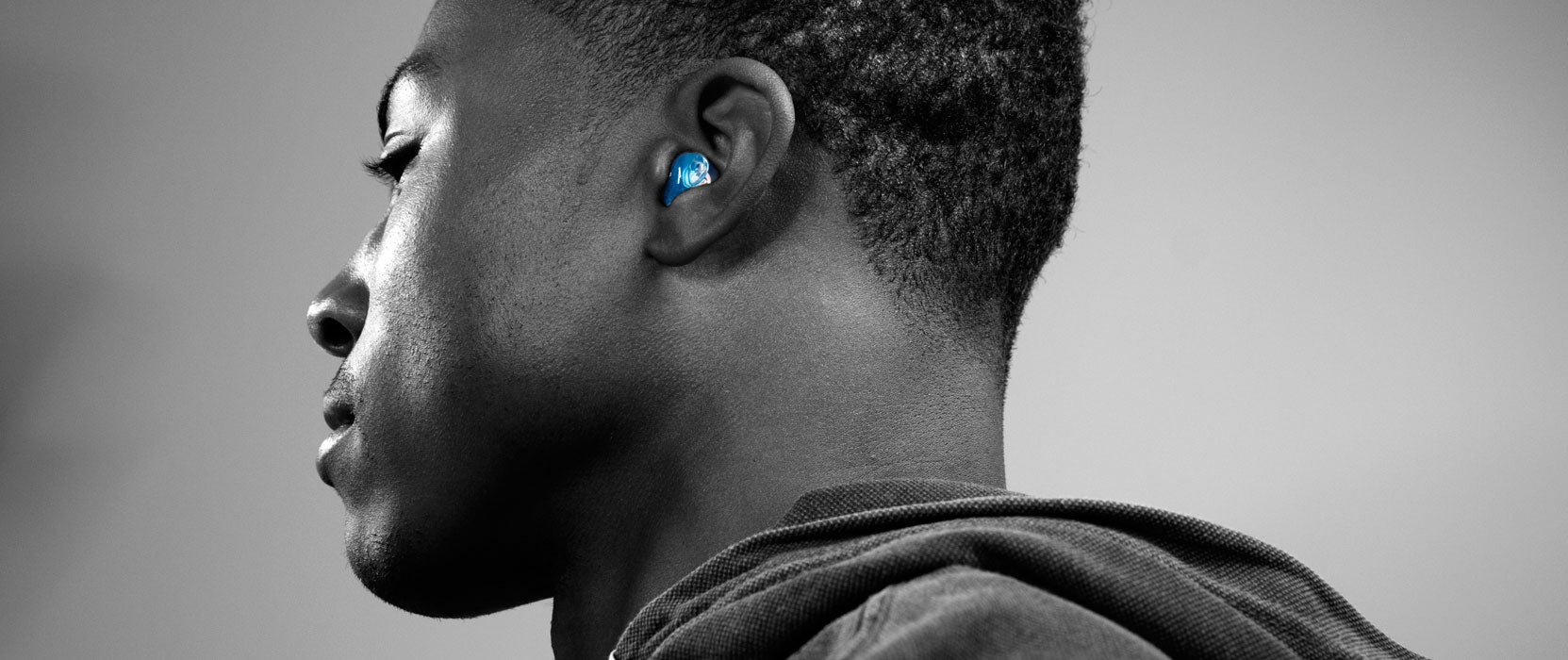 Close-up profile shot of a young man wearing a blue wireless earbud, with a focus on his ear and the detailed texture of his skin and haircut. the background is a soft, neutral gray.