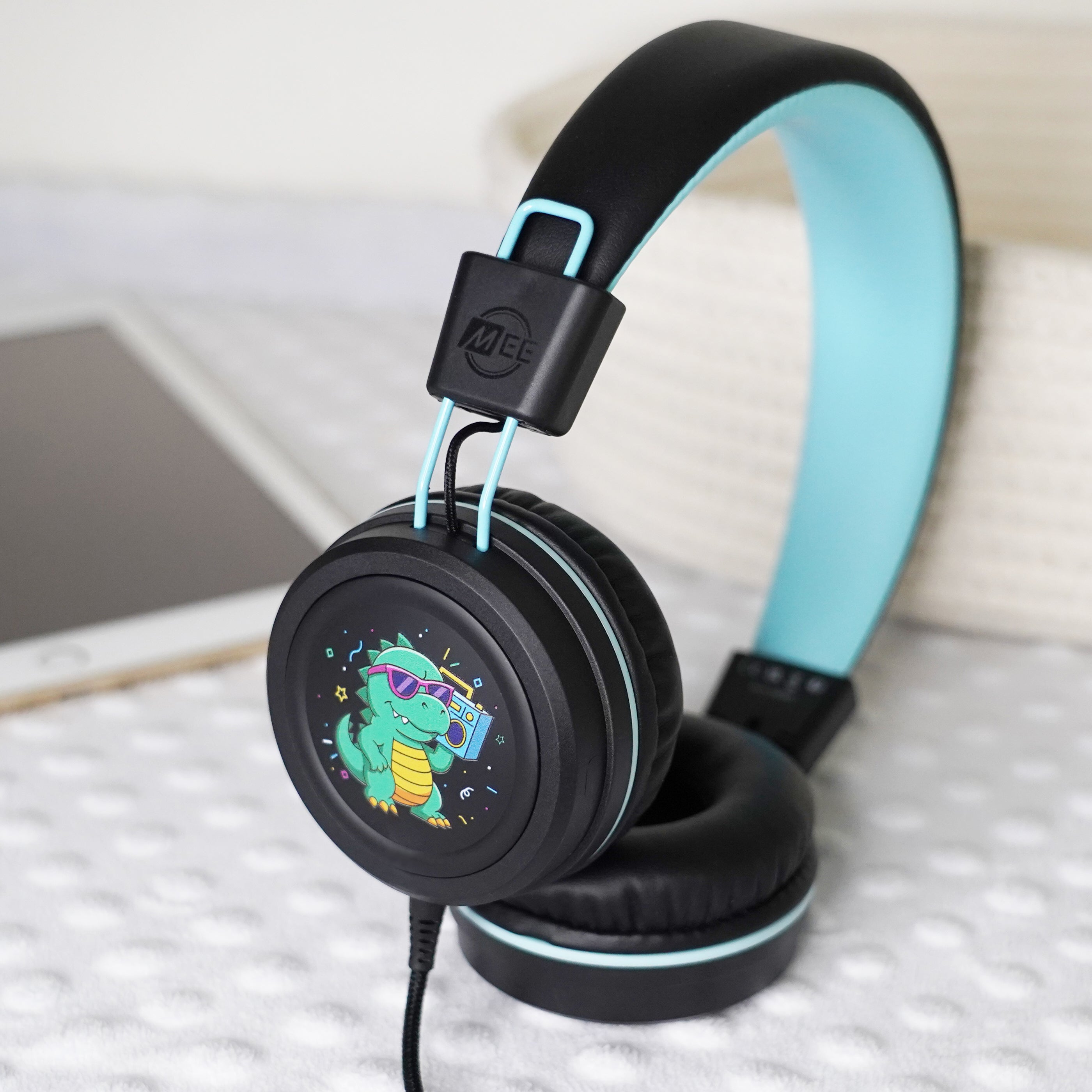 Black KidJamz KJ55AUX headphones with a cartoon dinosaur design holding a speaker in color faceplate on a light gray background