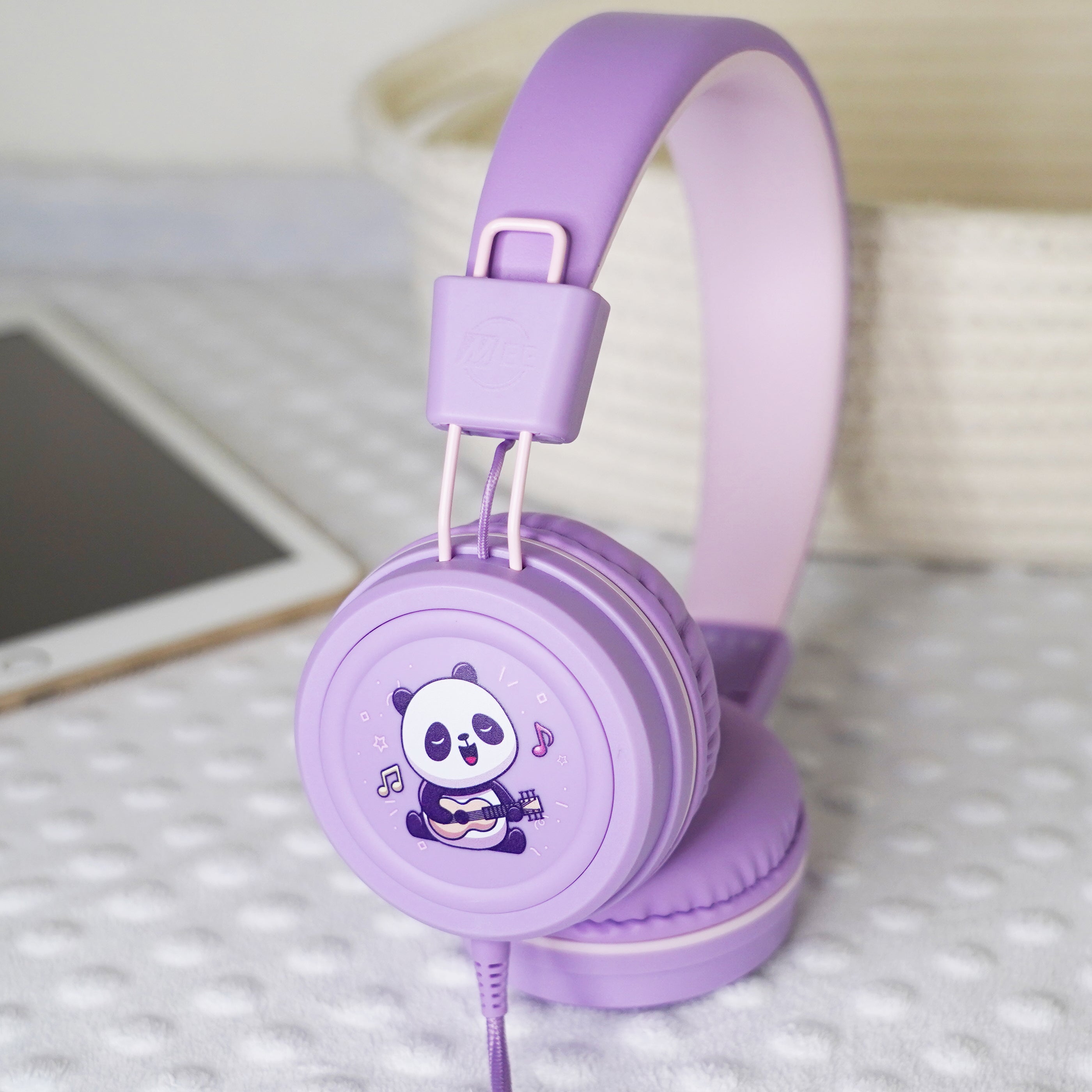 Lavender KidJamz KJ55AUX headphones with a cartoon Panda design holding a guitar while in color next to a tablet.