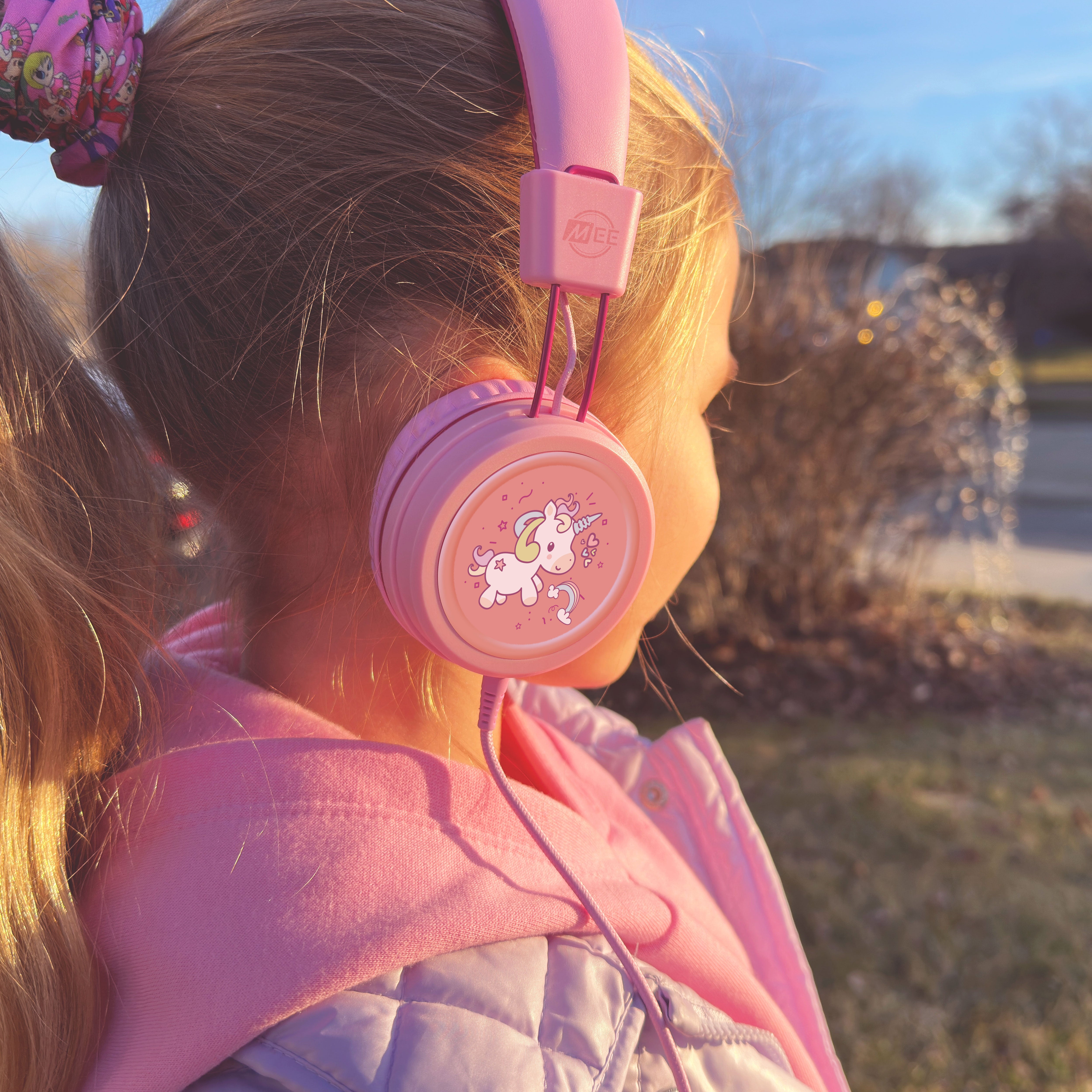 Young girl wearing pink KJ55AUX headphones with a unicorn design outdoors.